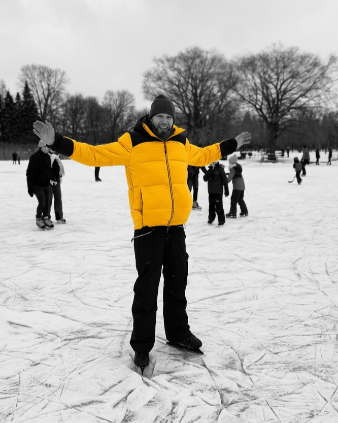 Man ice skating on a frozen pond, wearing a bright yellow jacket and a black beanie, with other skaters in the background and trees without leaves.