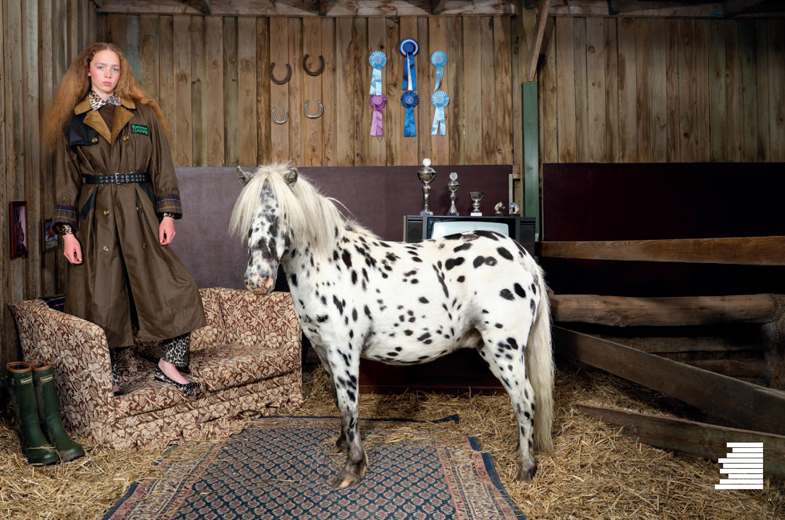 Image of a model in a Barbour Ganni collaboration jacket standing in a stable with a small, spotted Shetland pony