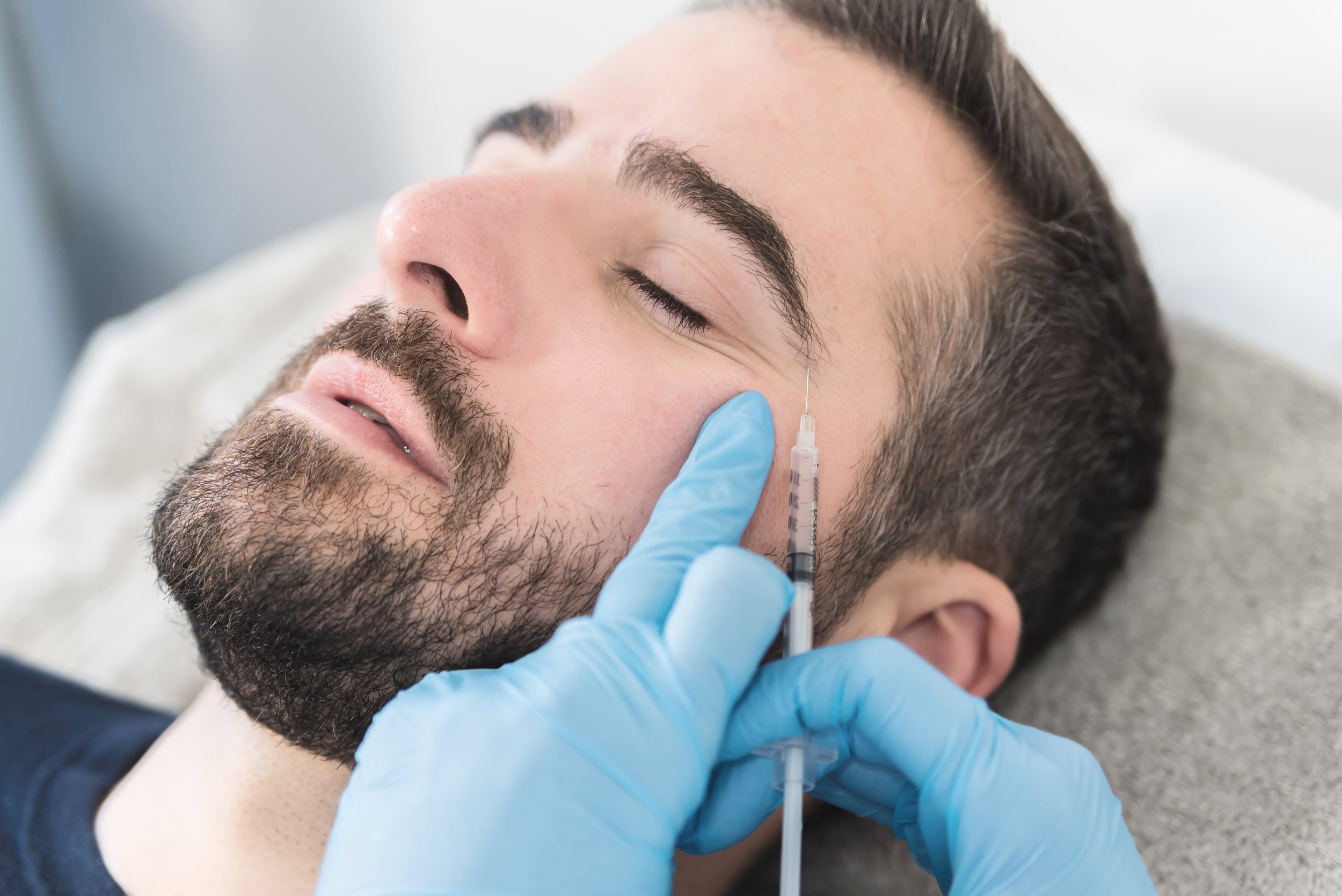 A man with a beard and closed eyes is lying down as a healthcare professional in blue gloves administers a cosmetic injection to his cheek.