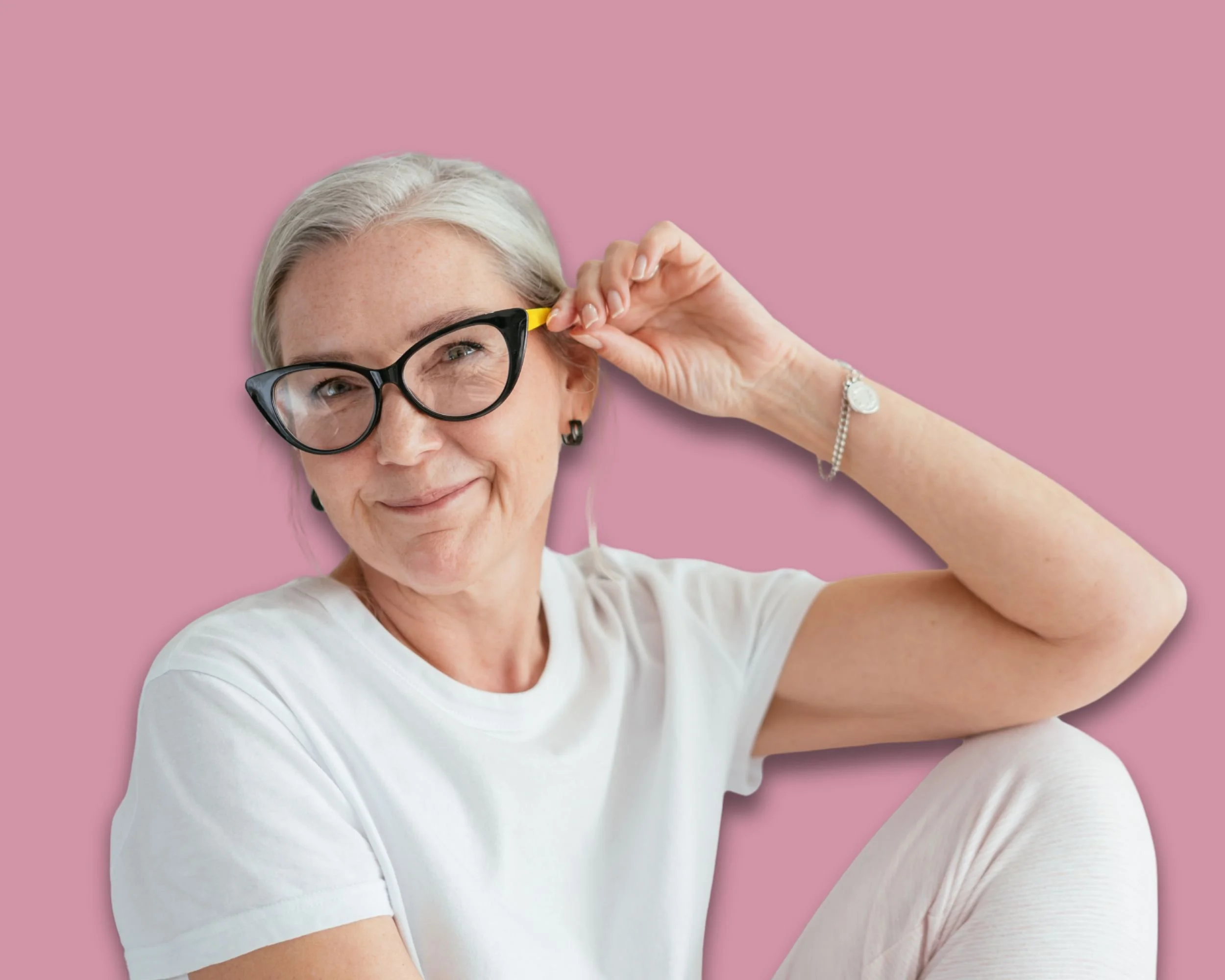 Eine lächelnde ältere Frau mit grauem Haar, Trägerin einer weißen T-Shirt, trägt große schwarze Brillen mit gelber Seite, sitzt vor rosa Hintergrund, hebt eine Hand zur Brille, trägt Armband und Ohrringe.