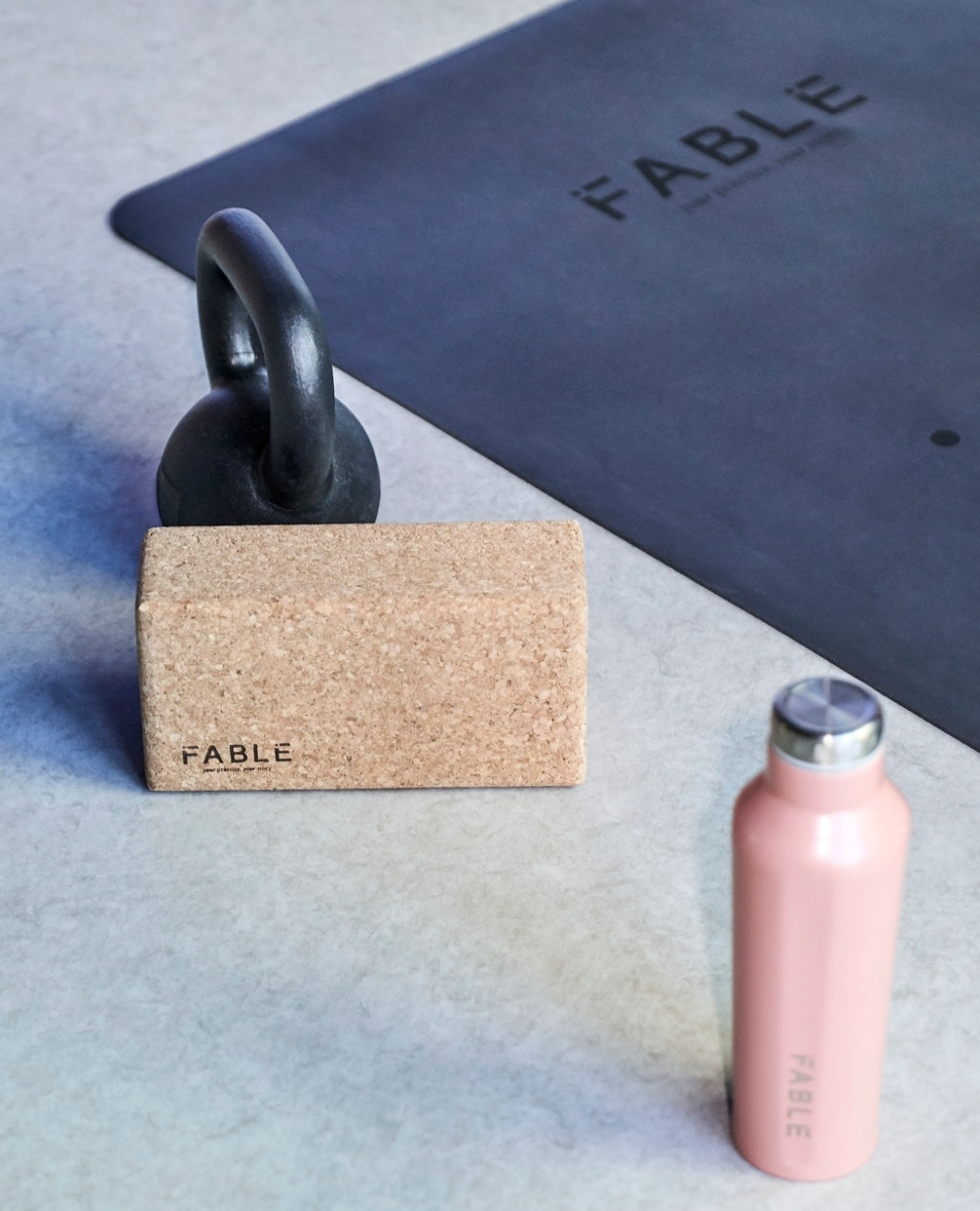 A black kettlebell, a cork block with the word 'FABLE' and the phrase 'your passions. your life.' on it, a black exercise mat with 'FABLE' written on it, and a pink reusable water bottle on a light-colored gym floor.