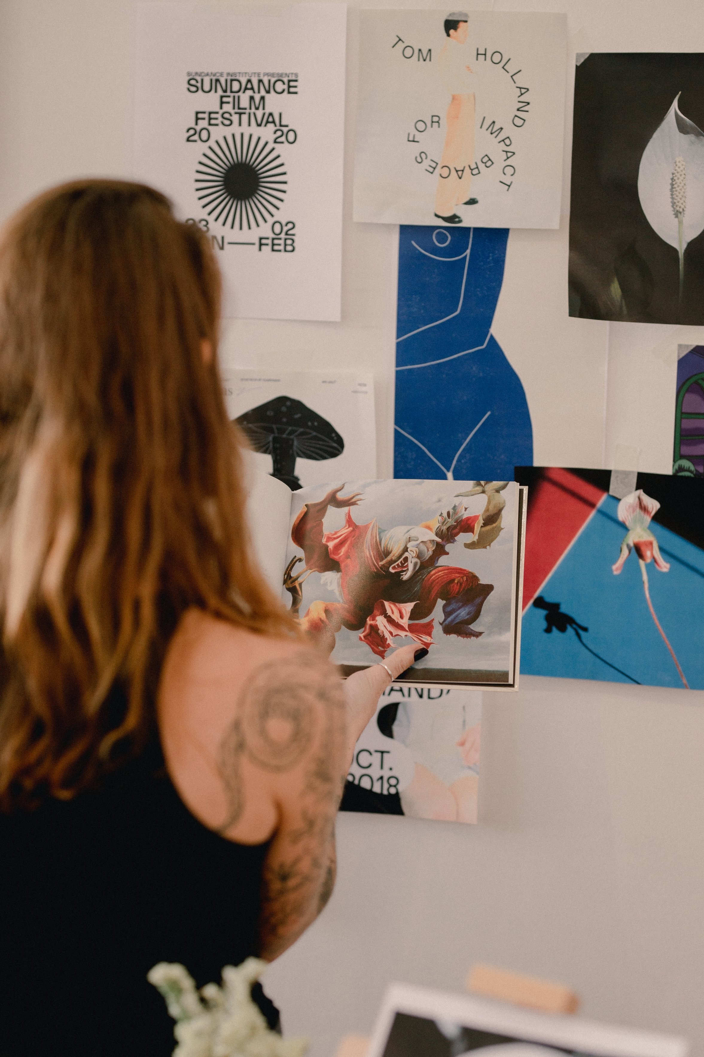 A woman with tattoos on her arm is pointing at a colorful picture of a lion on a wall decorated with various posters and artworks, including a film festival poster, a diagram of a face, a flower, and abstract art.