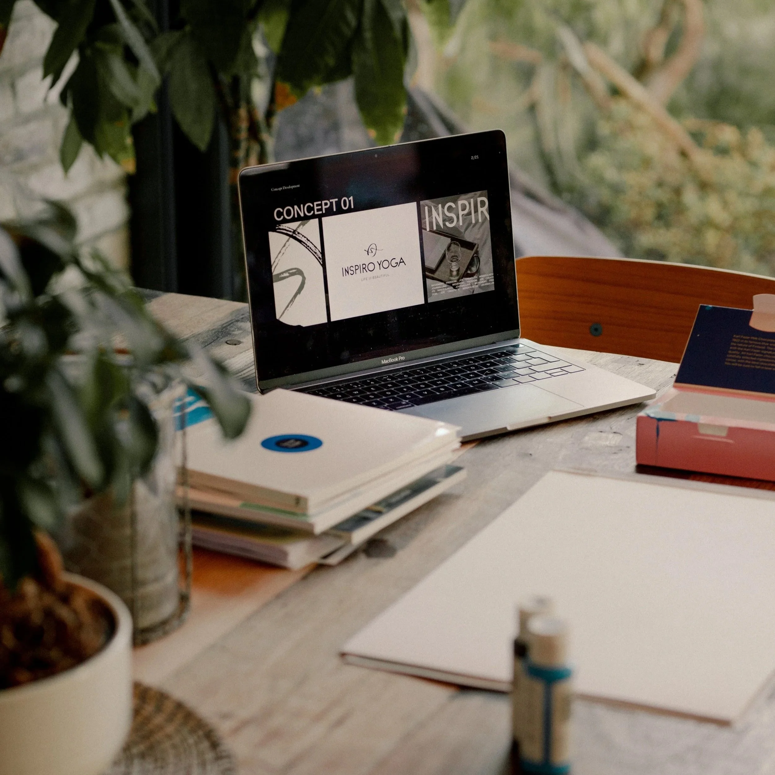 A laptop on a wooden desk displaying a presentation slide titled 'CONCEPT 01' with 'INSPIRE YOGA' and 'LIVE BEAUTIFULLY'. There are notebooks, a pink box, a white sheet, a plant in a pot, and a bottle of glue on the desk, with a window and greenery i