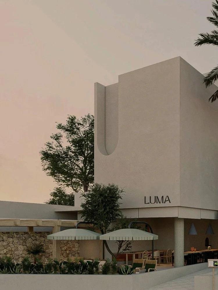 Modern white building with the word "LUMA" on it, outdoors seating area with umbrellas, trees, and a stone wall in the background during dusk or dawn.