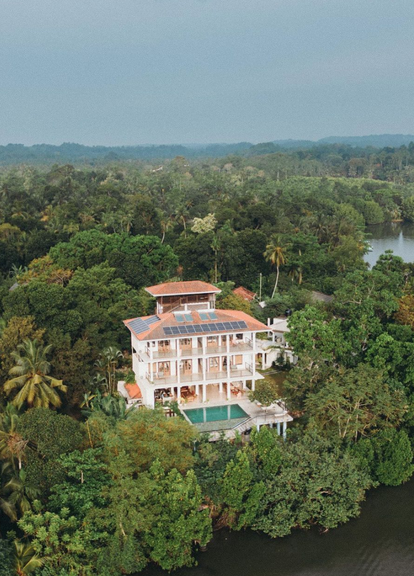 A large white house with multiple balconies and rooftop solar panels, surrounded by lush green trees, with a swimming pool in the backyard and water nearby.