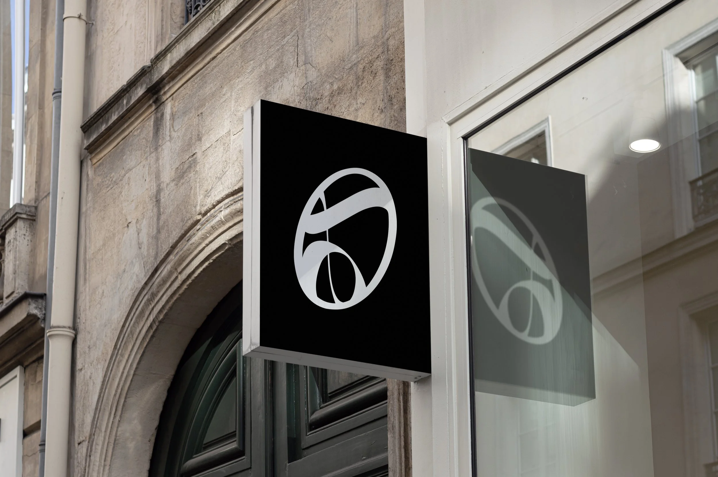 A black storefront sign with a white abstract logo mounted on an outdoor stone wall, with a reflection of the sign visible in a nearby glass window.
