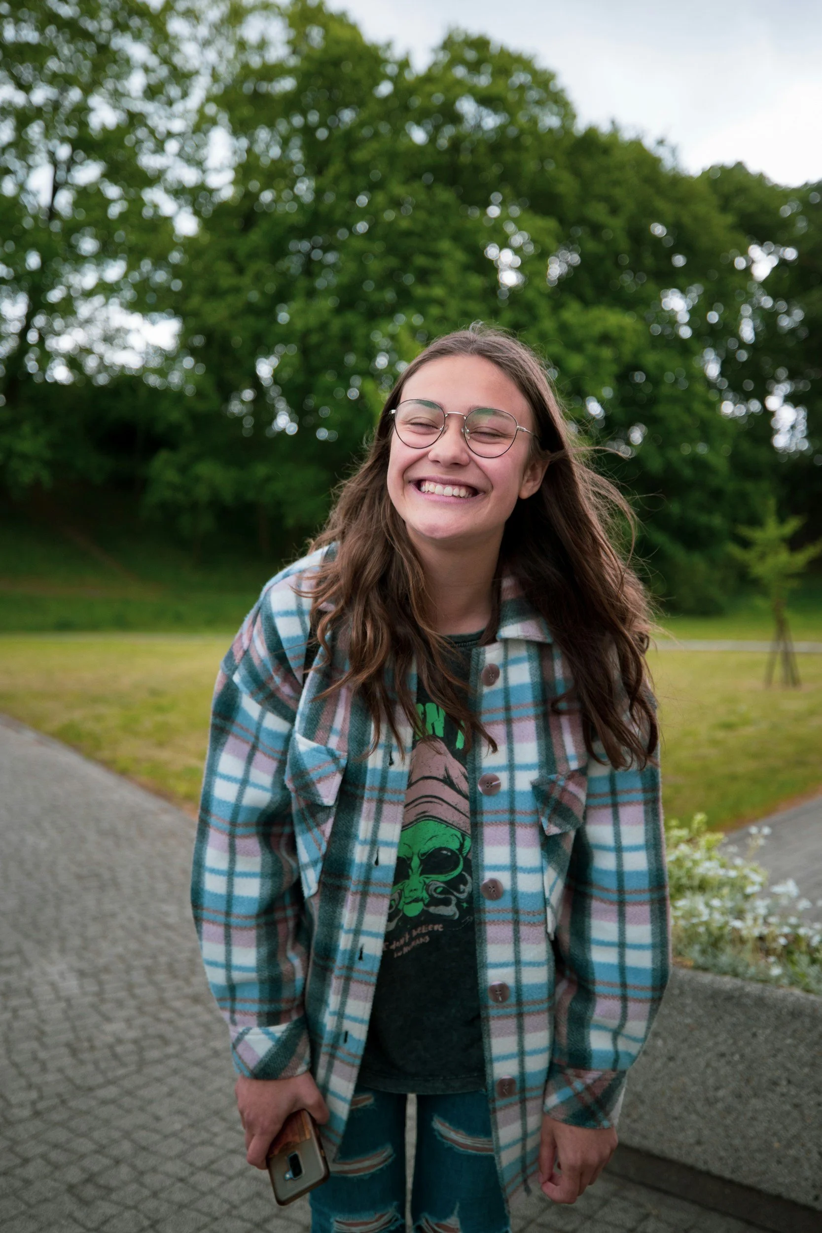 Young woman smiling and squinting outdoors, holding a phone, wearing glasses, a green graphic T-shirt, and a plaid jacket, standing on a paved sidewalk with green trees in the background.