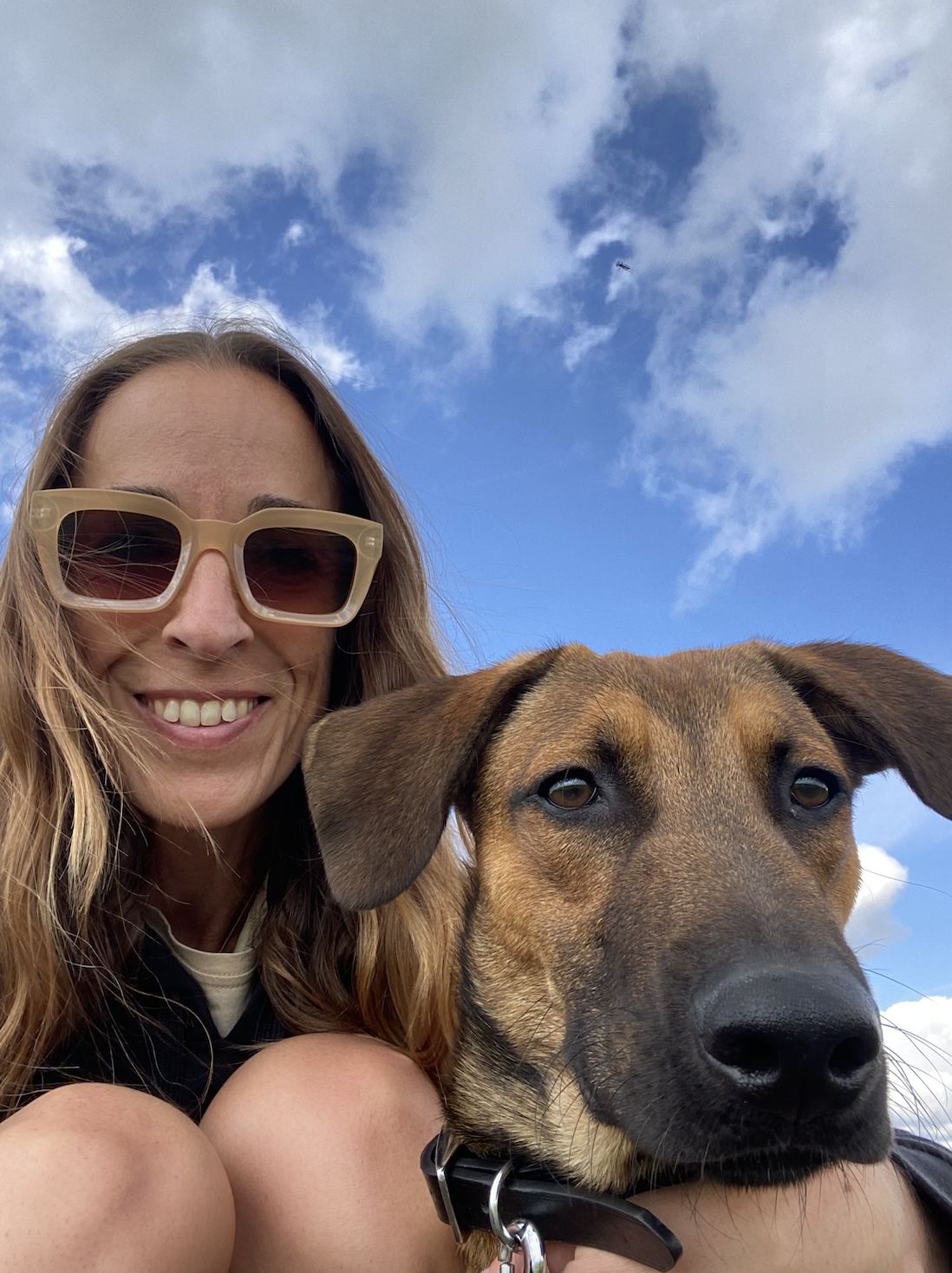 A woman with long, wavy hair wearing large, light-colored sunglasses smiling with a brown dog with floppy ears and dark eyes, against a blue sky with white clouds.