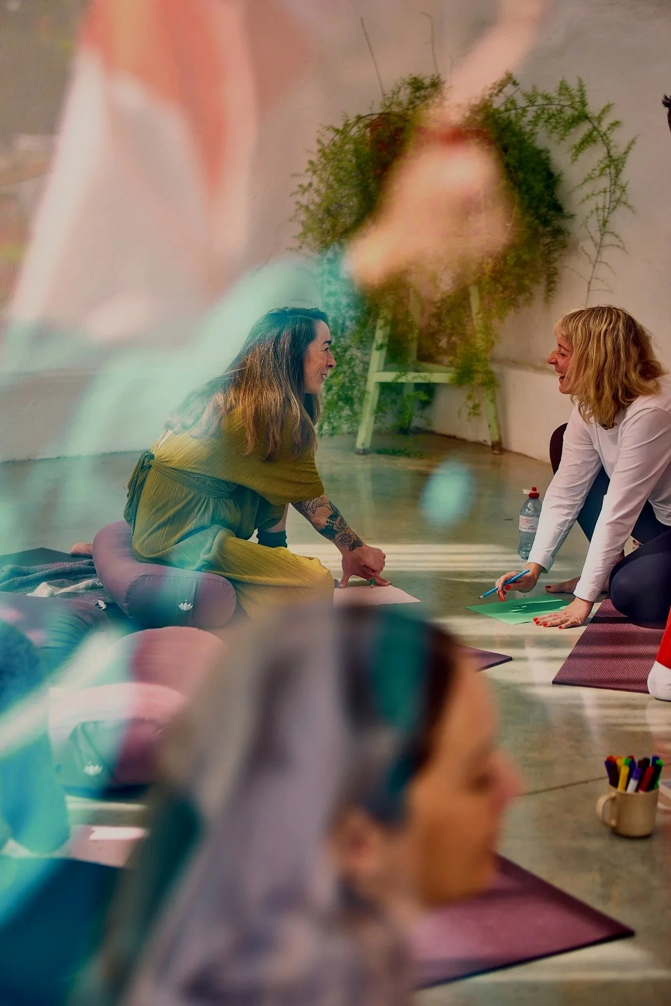 Two women in a bright room, smiling and engaging in a yoga or meditation session, with yoga mats, a water bottle, and a container of colored markers nearby, and a large blurred face projection on the wall in the background.