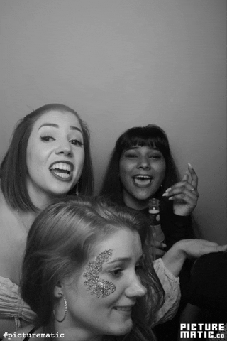 Three young women taking a selfie together indoors, one with face glitter, smiling and making a peace sign.