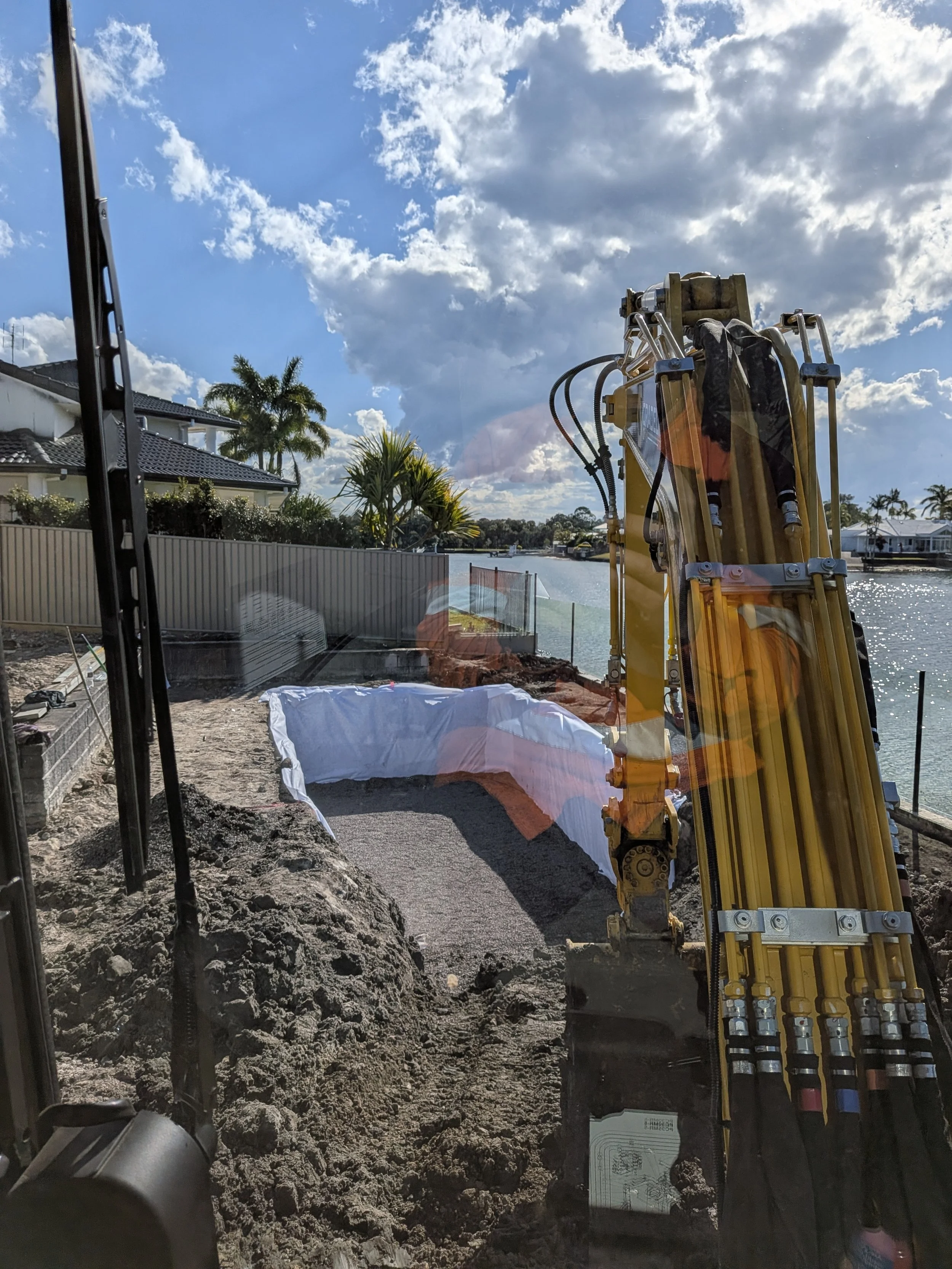 View of a construction site with a hydraulic excavator and a body of water in the background. The site includes dirt, construction materials, and landscaping with palm trees, under a partly cloudy sky.