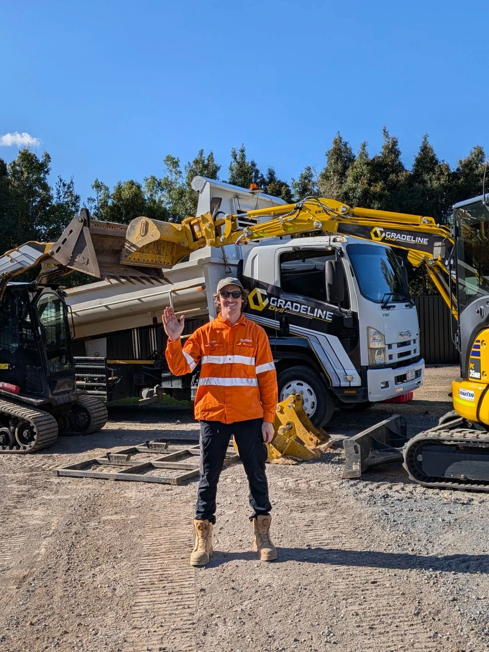 A man wearing beige work boots, dark pants, an orange jacket, and glasses stands outdoors, waving at the camera with a smile. He is in front of construction vehicles and equipment, including a large white truck with a yellow crane and other machinery, with trees and a blue sky in the background.