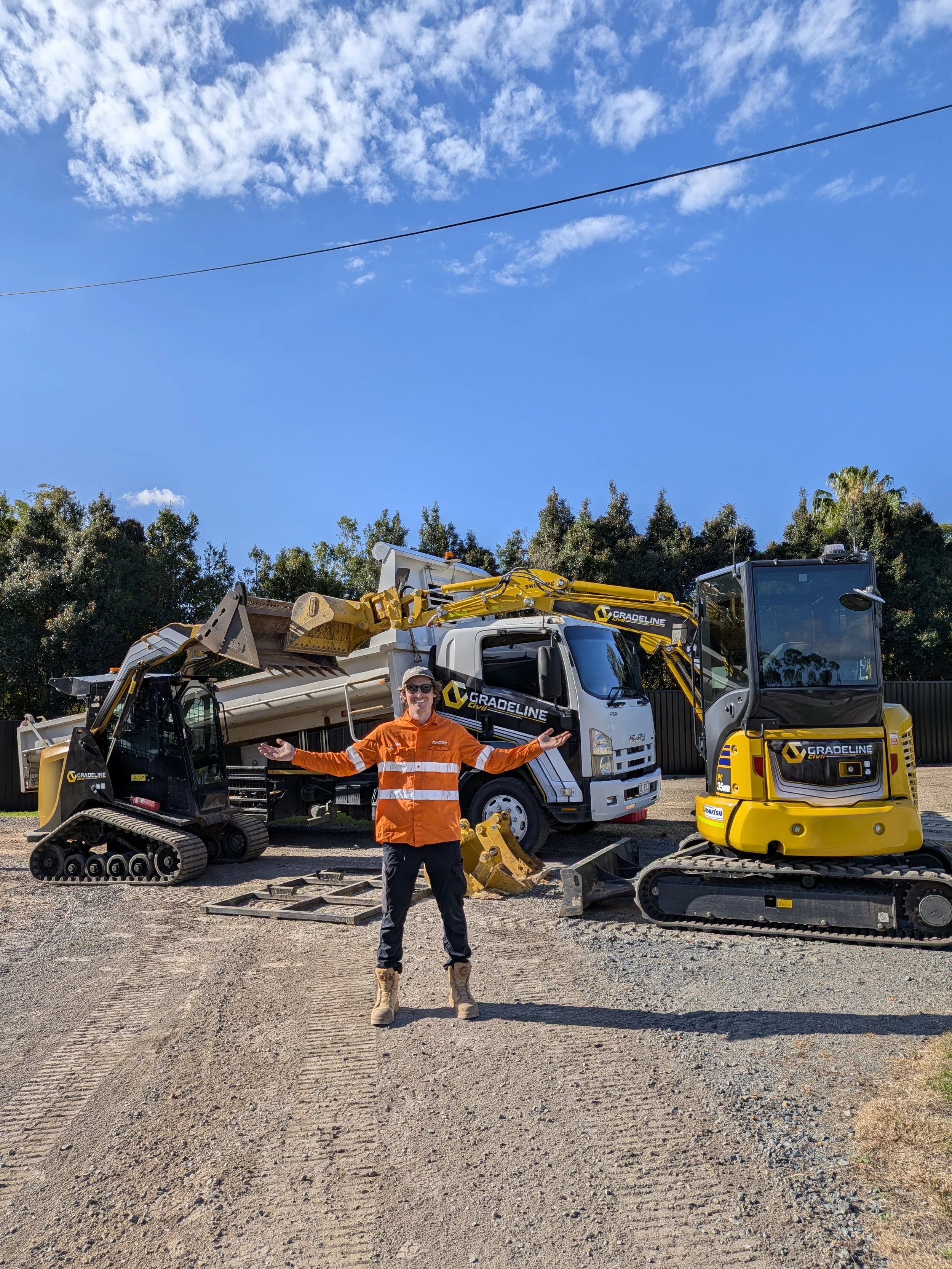 A person wearing an orange safety jacket and boots standing on a dirt lot with construction equipment, children, a truck, a skid steer loader, and a bright blue sky with clouds in the background.