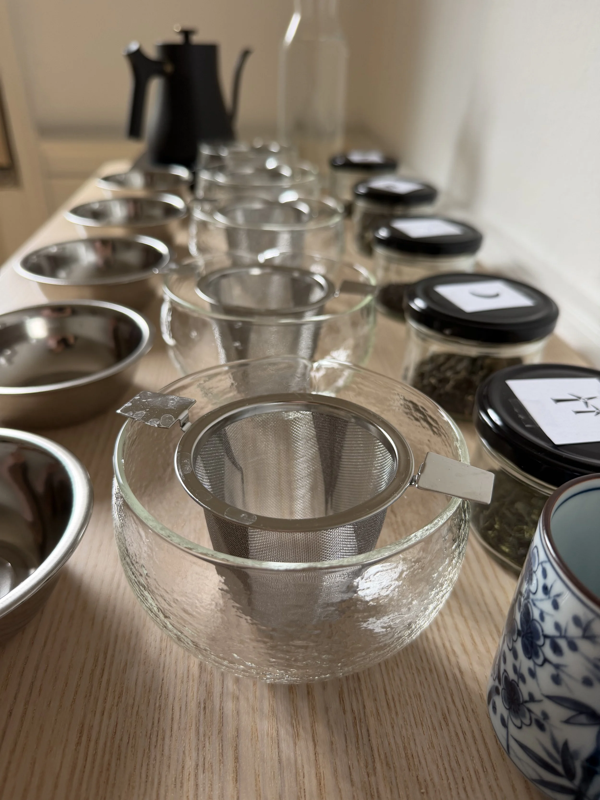 A glass tea strainer placed inside a clear glass bowl, with several small jars with black lids and white labels on the right, and a blue and white patterned teapot on the bottom right, all on a light wooden surface.