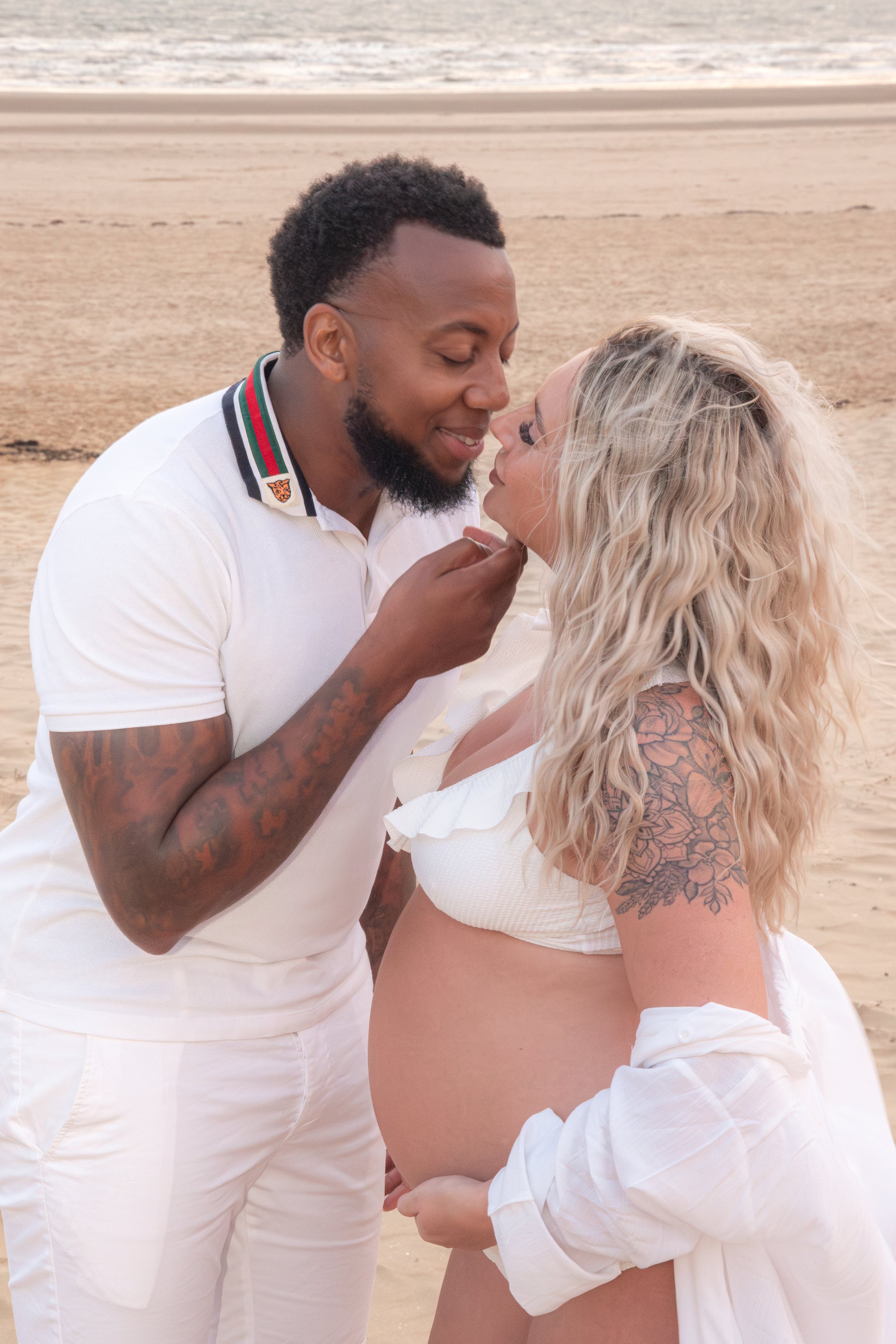 A couple embracing on a beach, with the woman showing her pregnant belly. They are wearing white clothing, and the man is gently holding the woman’s chin. The background features sand and the ocean.