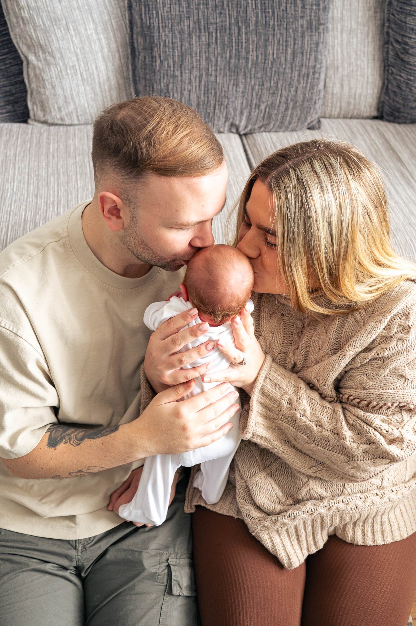 Couple holding and kissing a newborn baby sitting on a gray sofa.