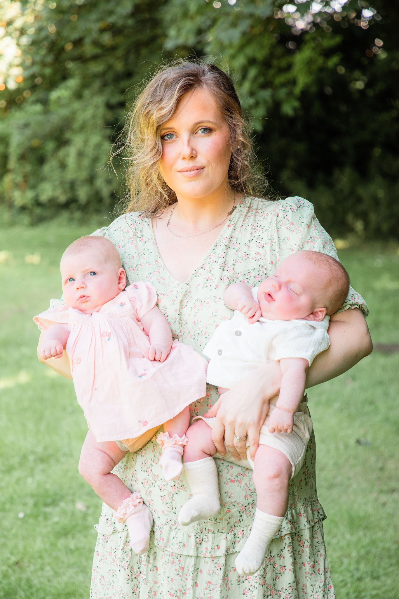 Woman holding two babies outdoors, wearing floral dress.