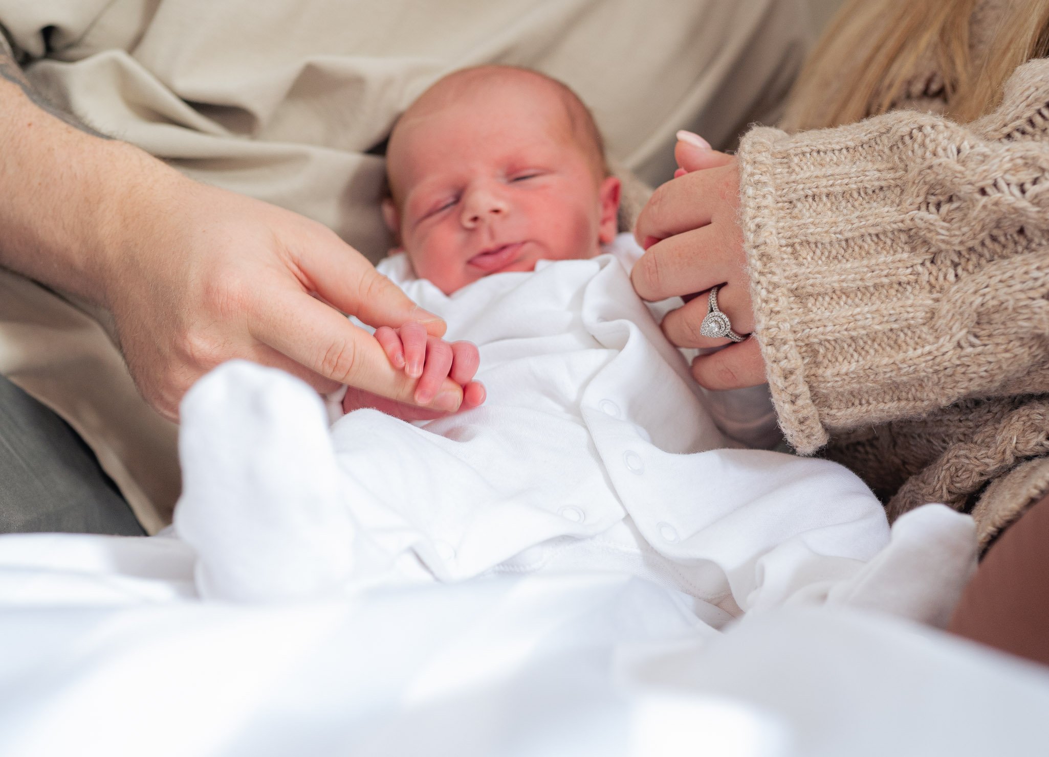 Newborn baby held by parent's hands, wearing white clothes and wrapped in a cozy blanket. Parent wearing a knitted sweater and a ring.