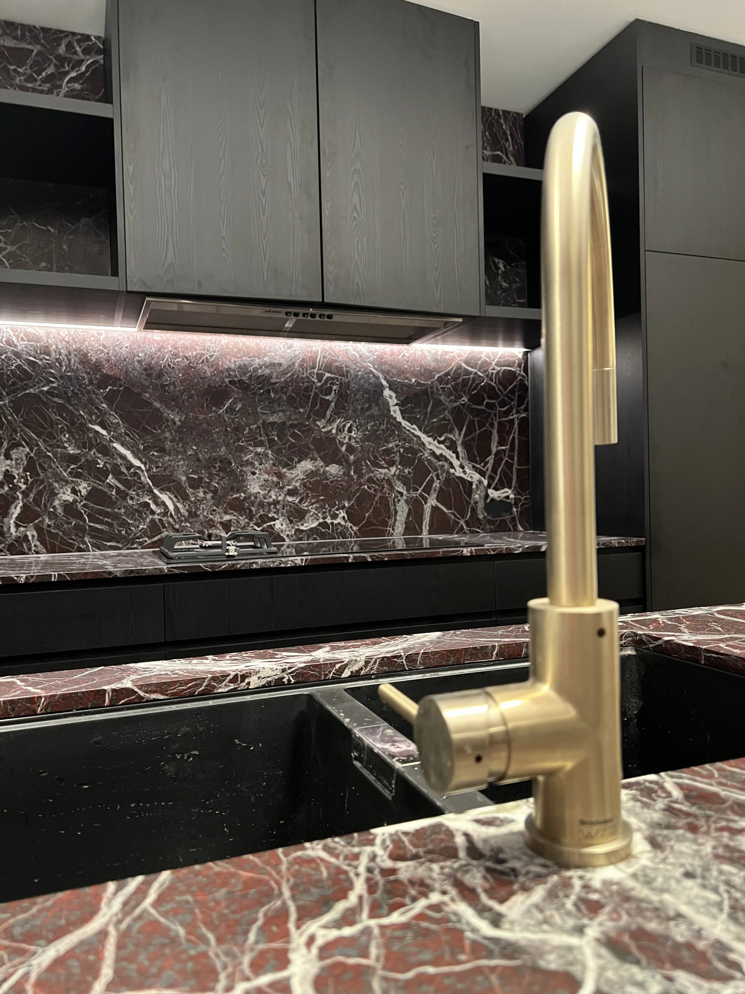 Close-up of a modern kitchen sink with a brass faucet, red and white marble countertop, black sink, gray cabinets, marble backsplash, and built-in stovetop in the background.