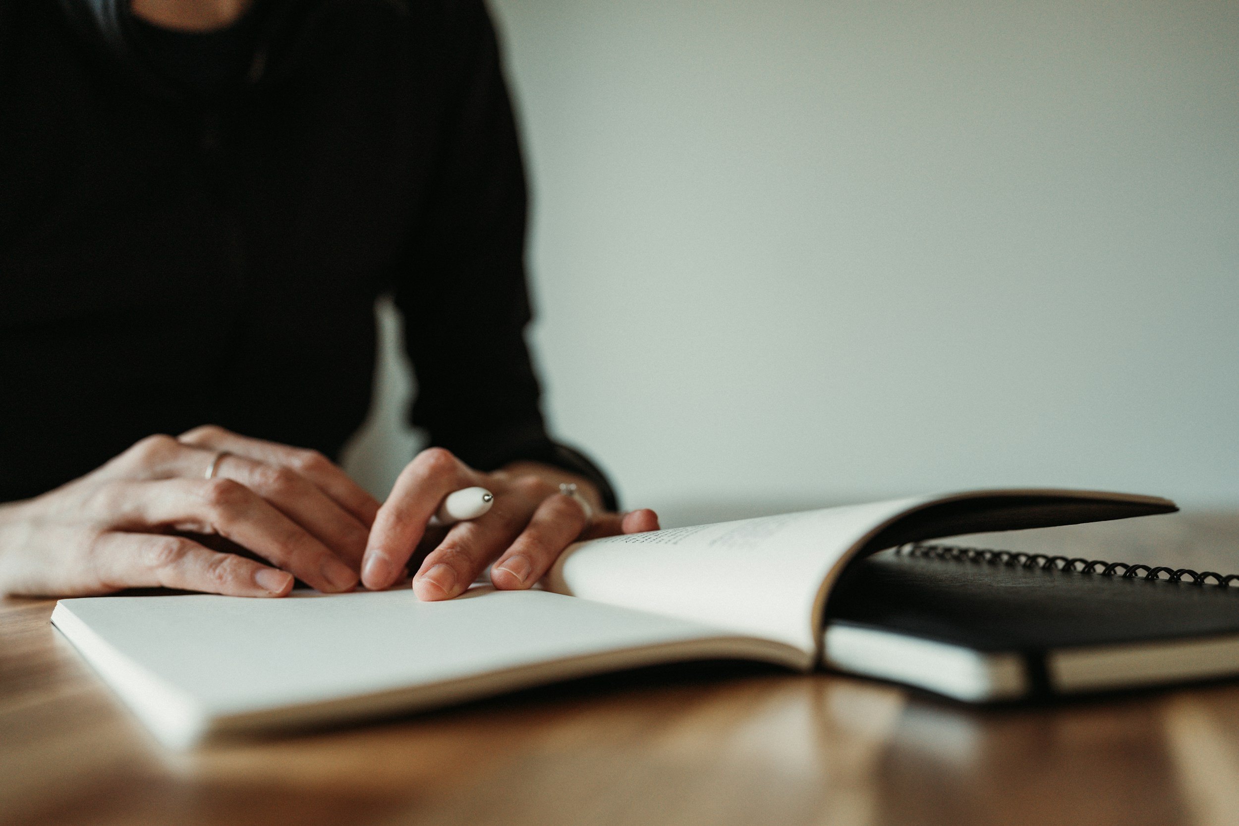 Person wearing black long sleeve shirt writing in a notebook with a white pen at a wooden table.