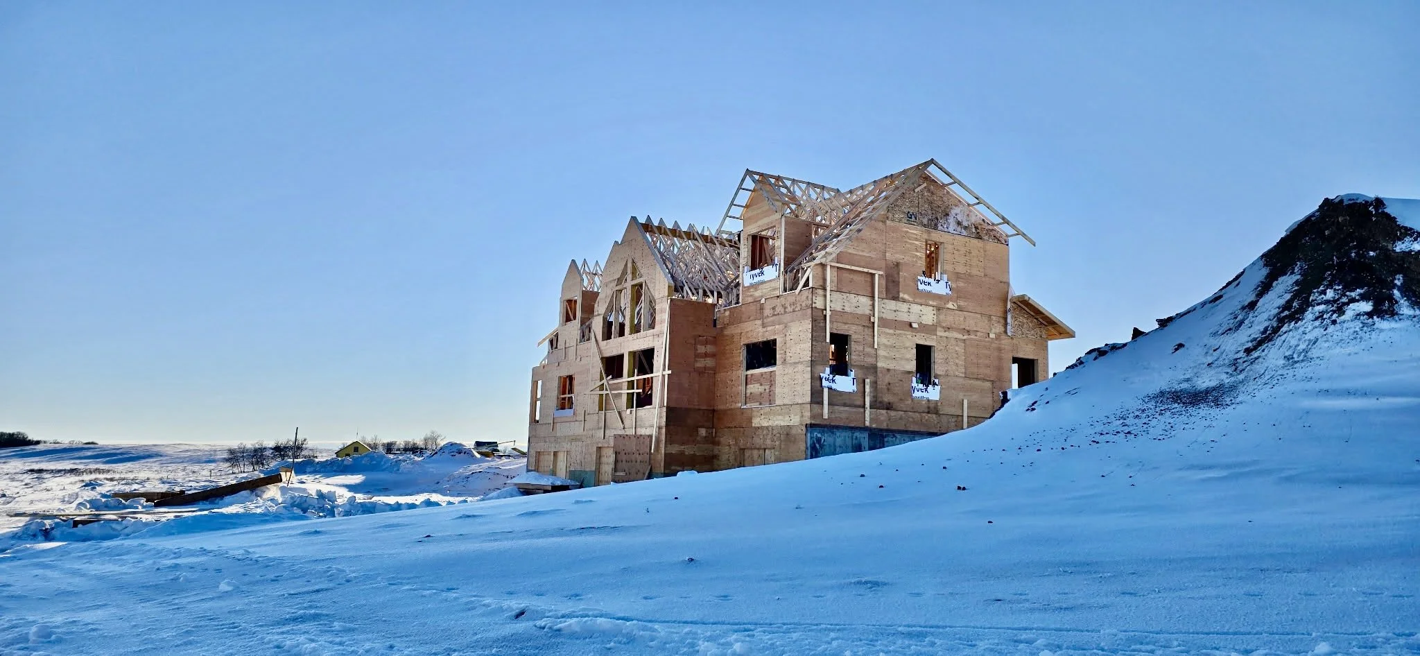 Construction site with wooden framing and open window and door frames, over a flat landscape with a cloudy sky.