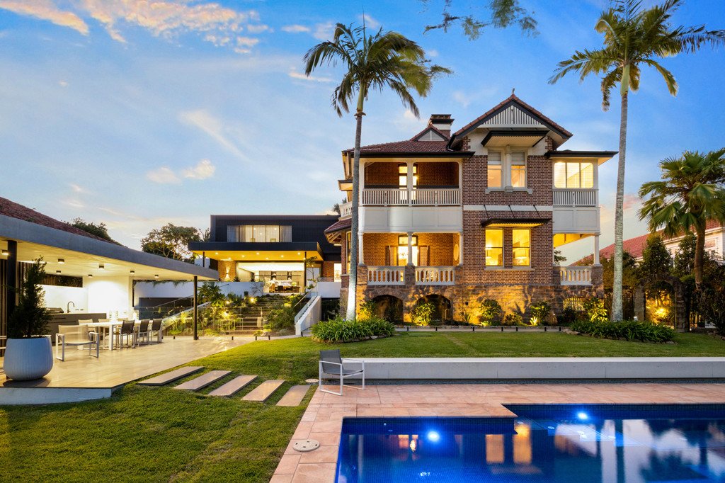 A luxurious multi-story house with a brick exterior, large windows, and a spacious balcony, surrounded by palm trees and landscaped gardens, with a swimming pool in the foreground during the evening.