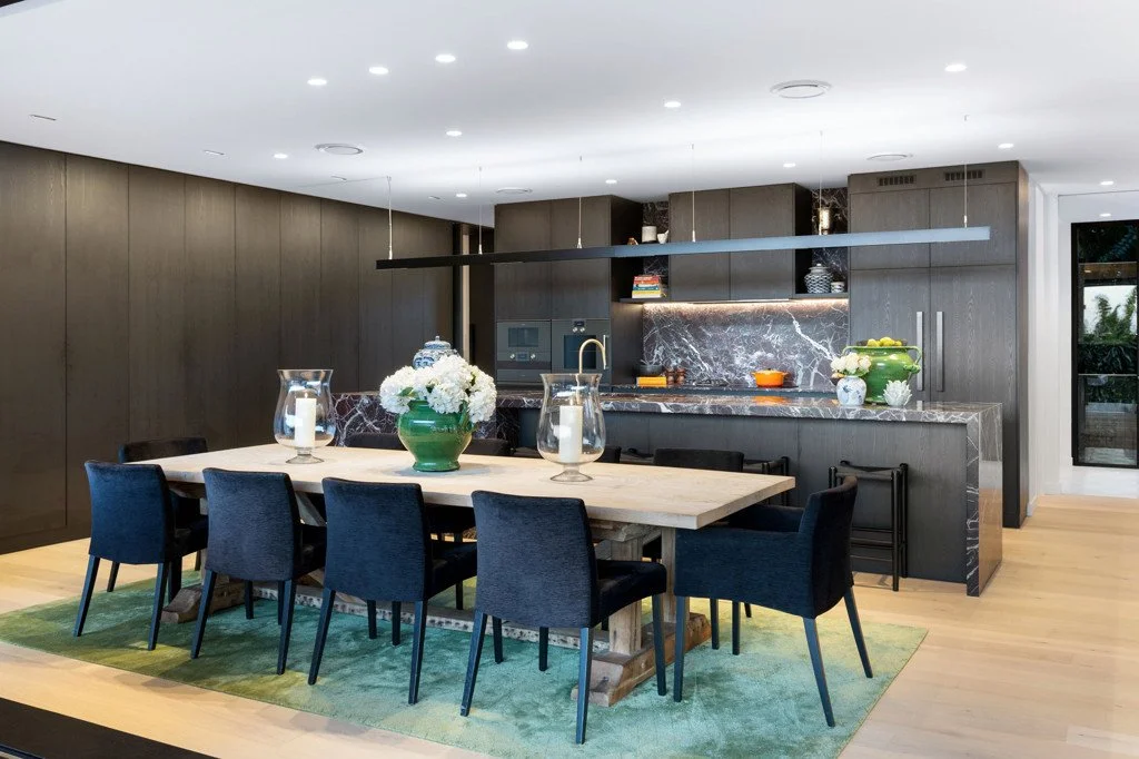 Modern open-plan kitchen and dining area with a large wooden table, eight black chairs, green rug, and sleek dark cabinetry with marble backsplash.