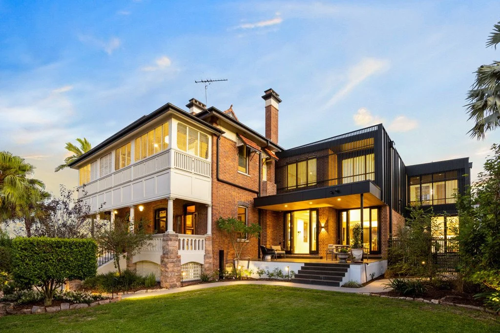 A two-story house with brick exterior, modern black additions, and large windows, surrounded by greenery and a well-maintained lawn under a blue sky.