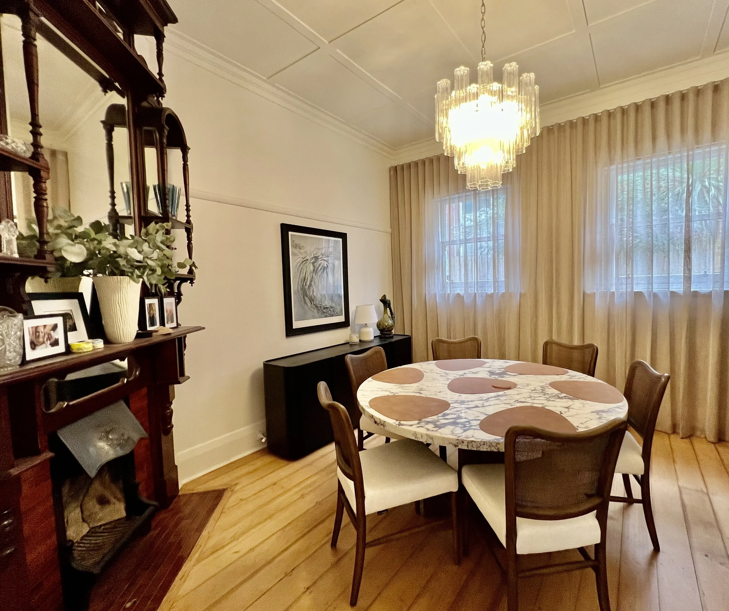 Modern living room with high vaulted ceilings, white walls, a fireplace with a black marble surround, and beige sofas arranged in a U-shape around a central green ottoman. Large windows and glass doors open to a backyard garden. Decor includes a horse-themed artwork, a flat-screen TV, and various decorative vases and art pieces.