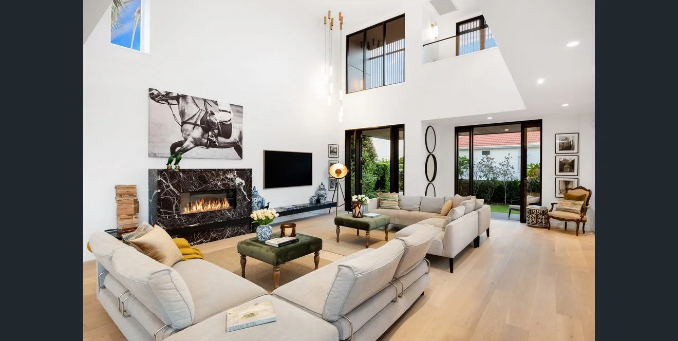 Modern living room with high vaulted ceilings, white walls, a fireplace with a black marble surround, and beige sofas arranged in a U-shape around a central green ottoman. Large windows and glass doors open to a backyard garden. Decor includes a horse-themed artwork, a flat-screen TV, and various decorative vases and art pieces.