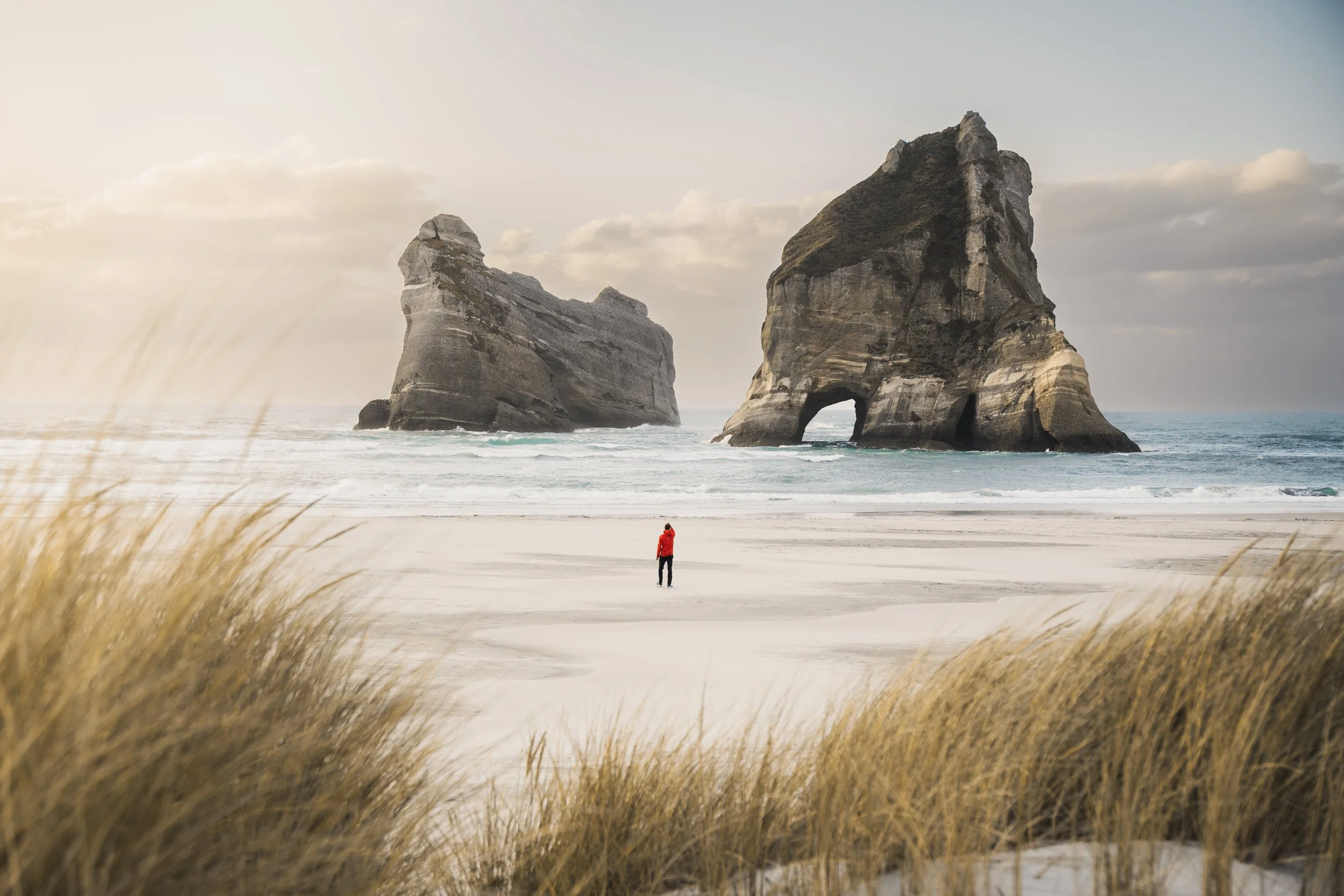 A person in a red jacket standing on a sandy beach near the ocean with two large rock formations in the background and dry grass in the foreground.