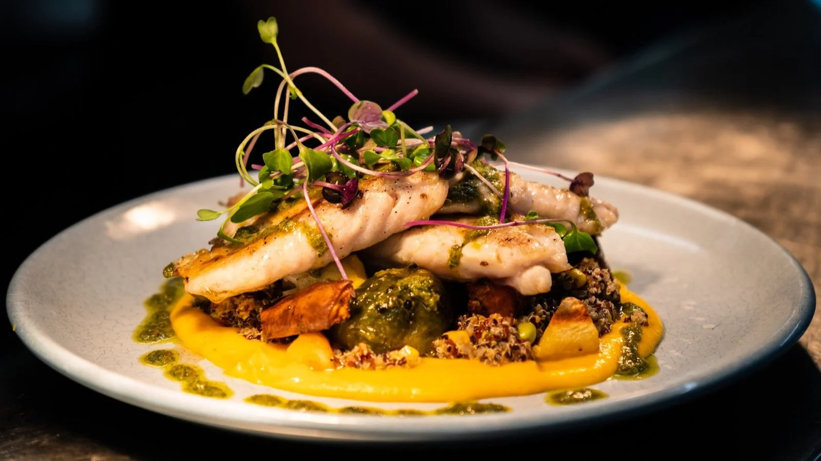 Gourmet dish with grilled fish, microgreens, green sauce, and colorful vegetables on a white plate