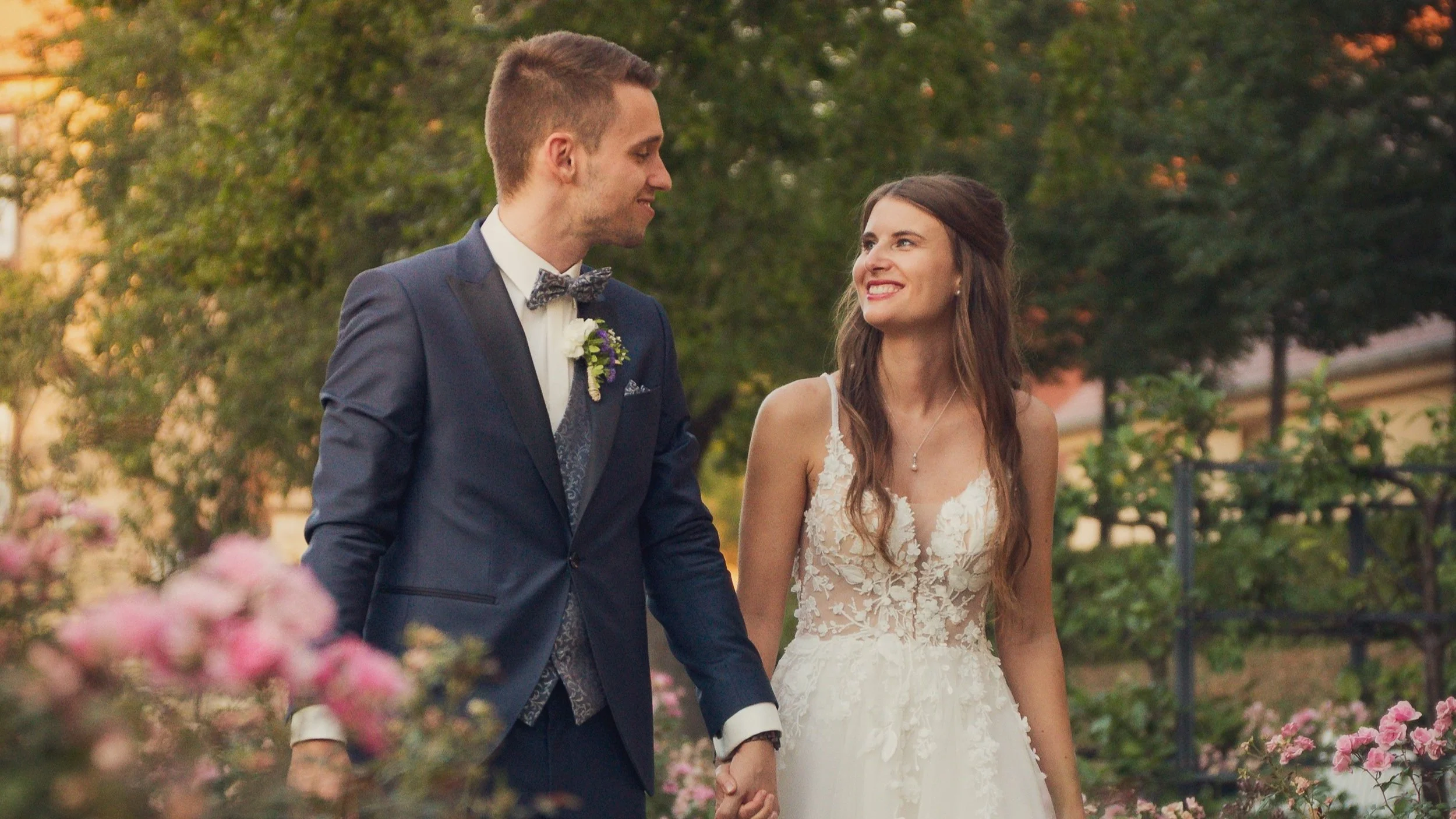 A bride and groom holding hands and smiling at each other outdoors during their wedding, with trees and pink flowers in the background.