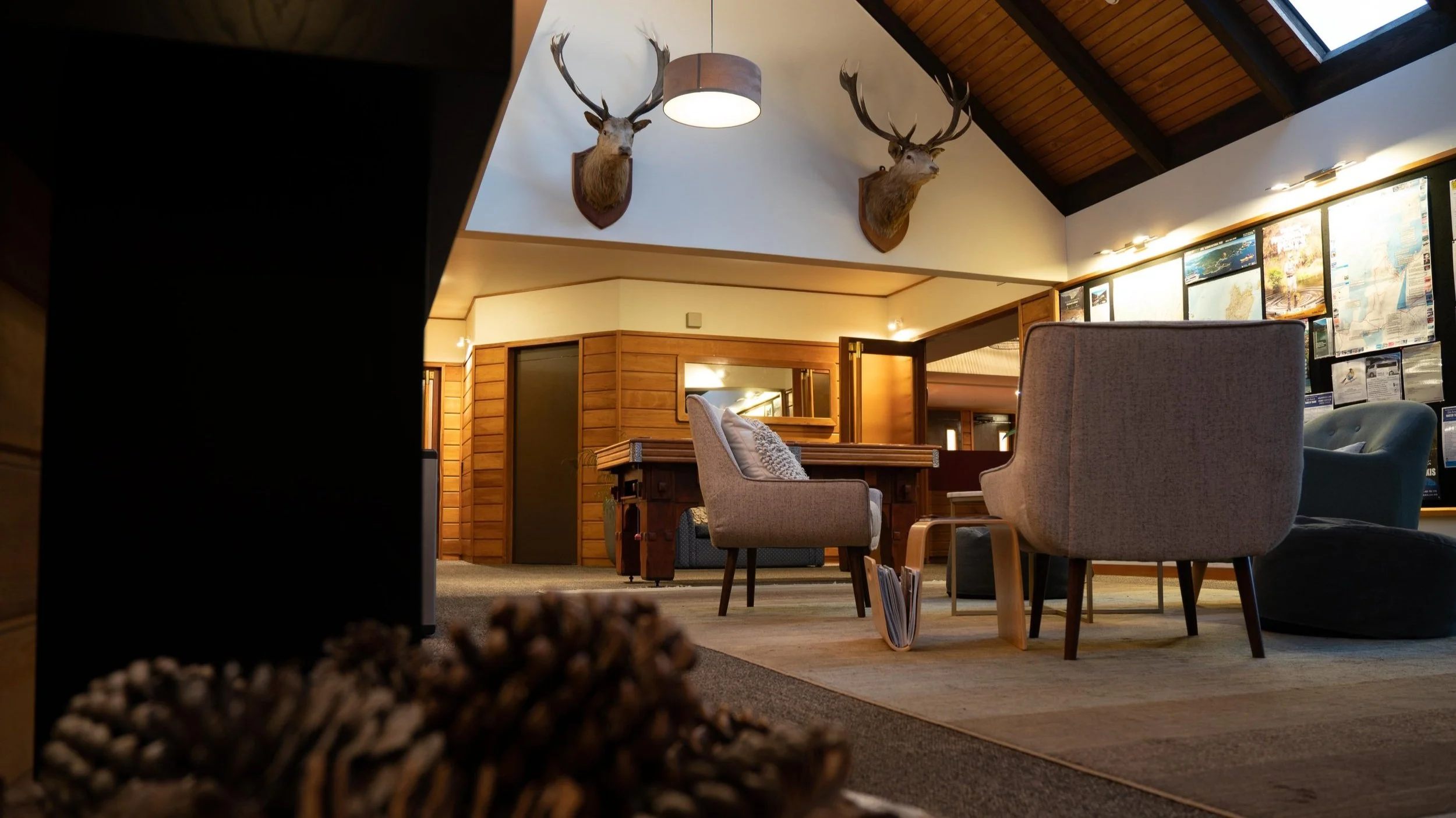 Inside a cozy lodge with wooden walls and ceiling, decorated with two mounted deer heads, furnished with armchairs, a table, and a magazine holder, and a wall display of maps and notices.