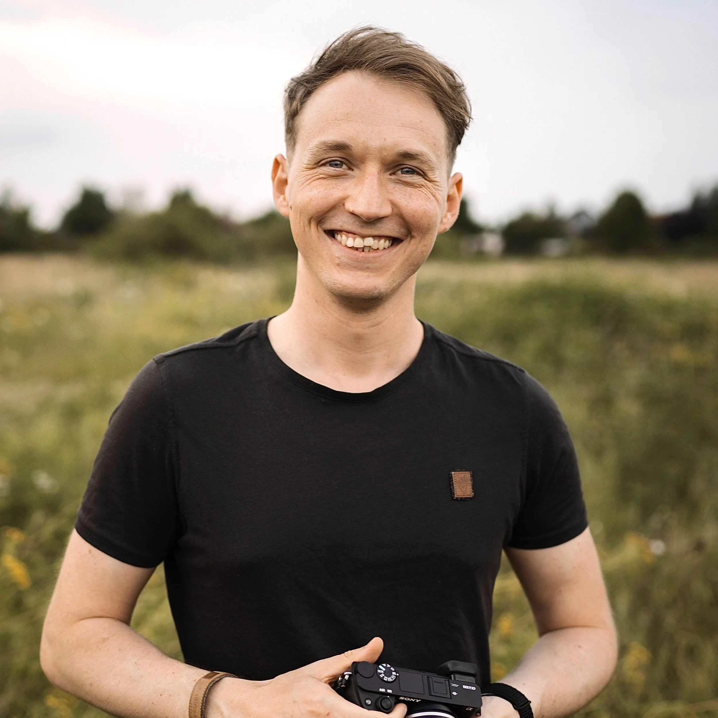 A smiling man outdoors holding a camera in a grassy field with trees in the background.