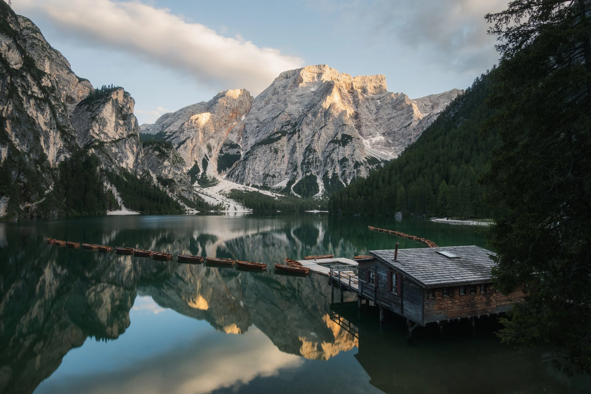 A tranquil mountain lake surrounded by tall rocky mountains and dense evergreen forest, with a boathouse and a line of small boats on the water.