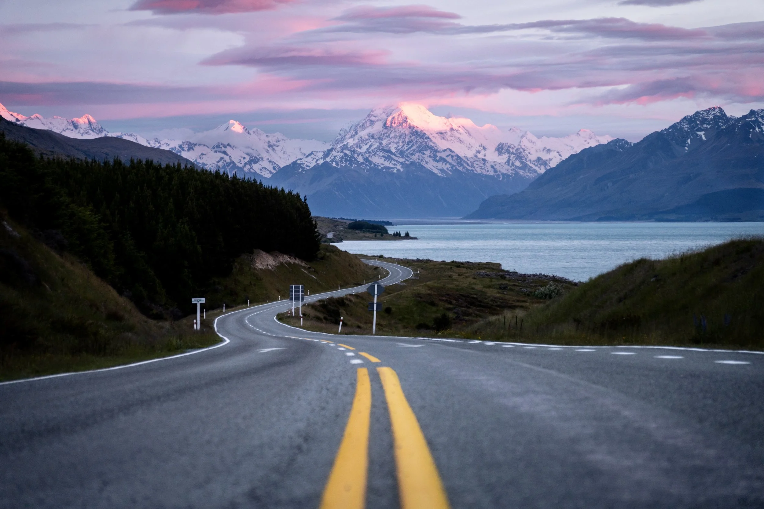 Winding mountain road leading towards snow-capped mountains near a lake during pink and purple sunset sky.