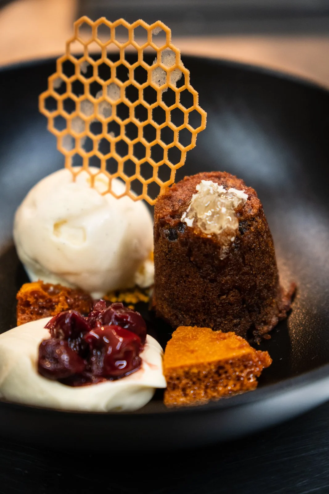 A dessert plate with a scoop of vanilla ice cream, a small slice of cake, a dollop of whipped cream with cherry topping, a mini Bundt cake, and a decorative caramel honeycomb piece on top.