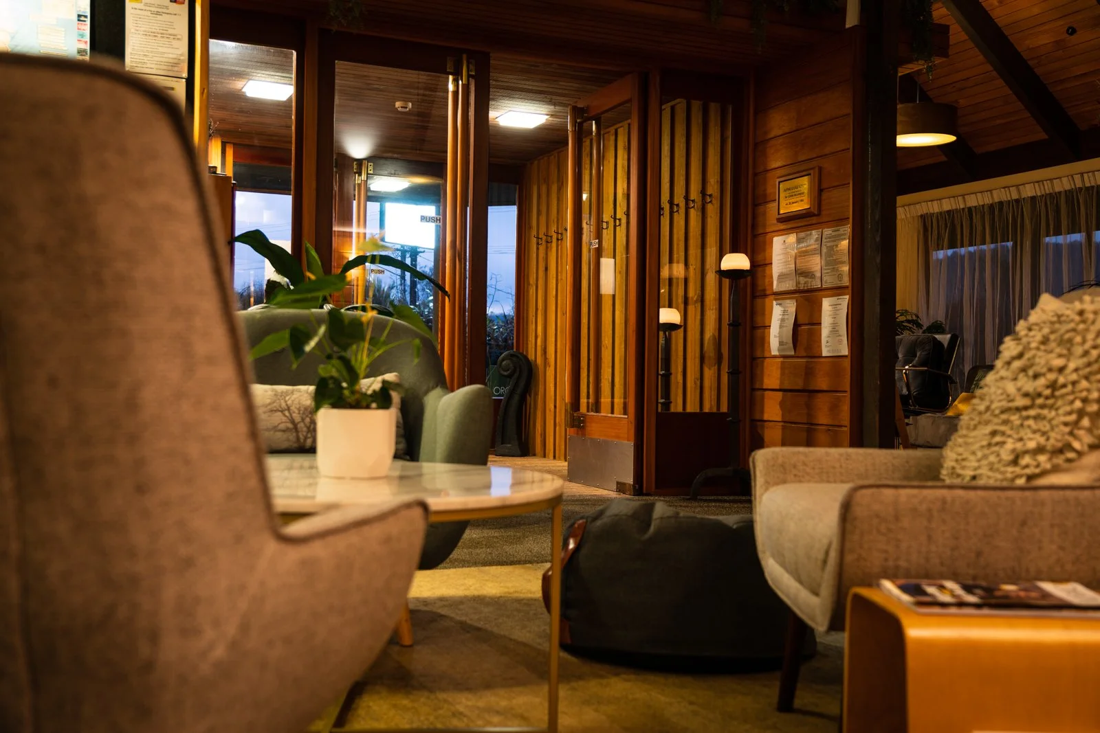 A cozy hotel lobby with wooden walls, a round table with a potted plant, surrounded by upholstered chairs and sofas, and a hallway with illuminated lamps and a partially open door leading outside.