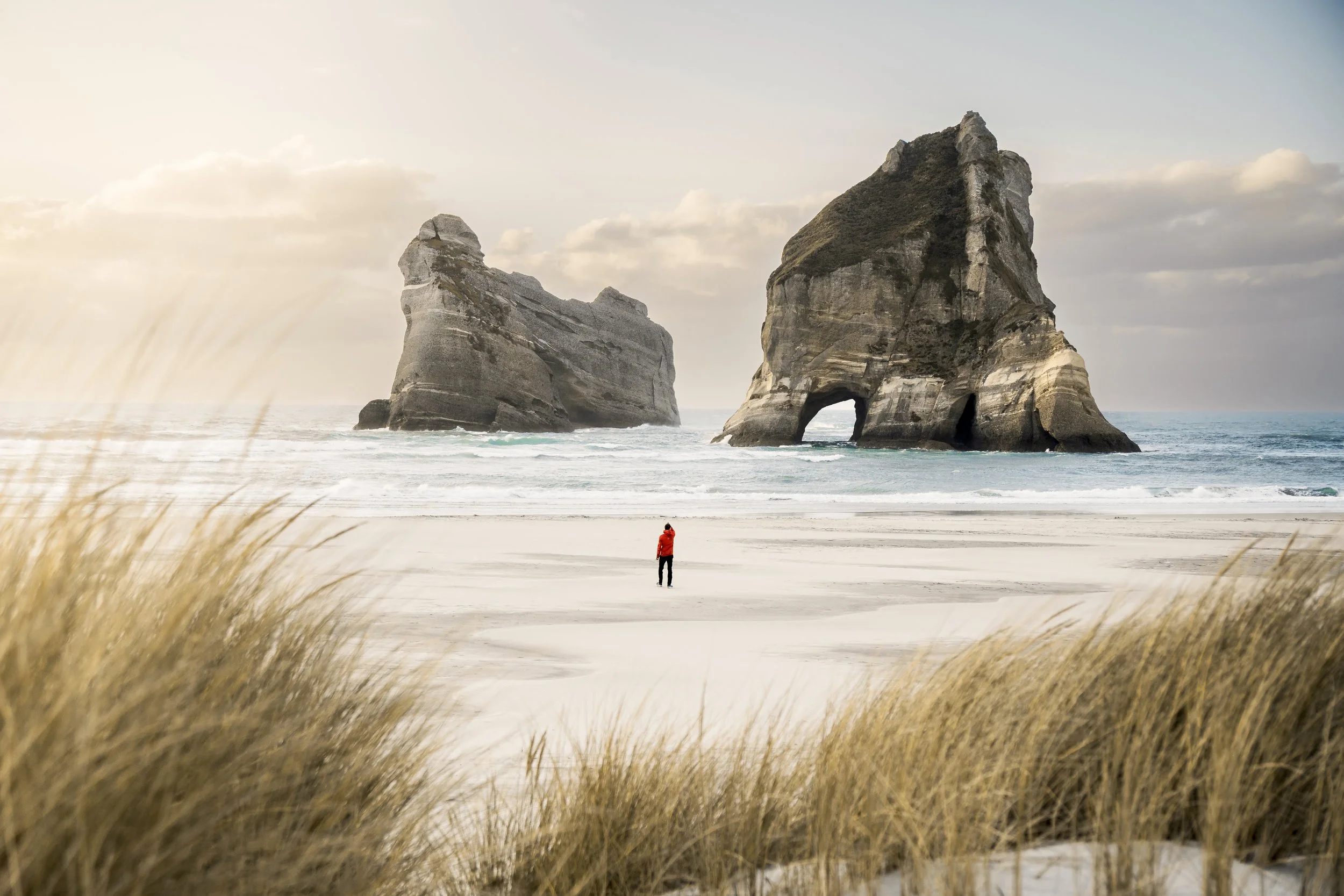 A person in a red jacket standing on a sandy beach with large rock formations in the ocean behind them, surrounded by grasses and cloudy sky.