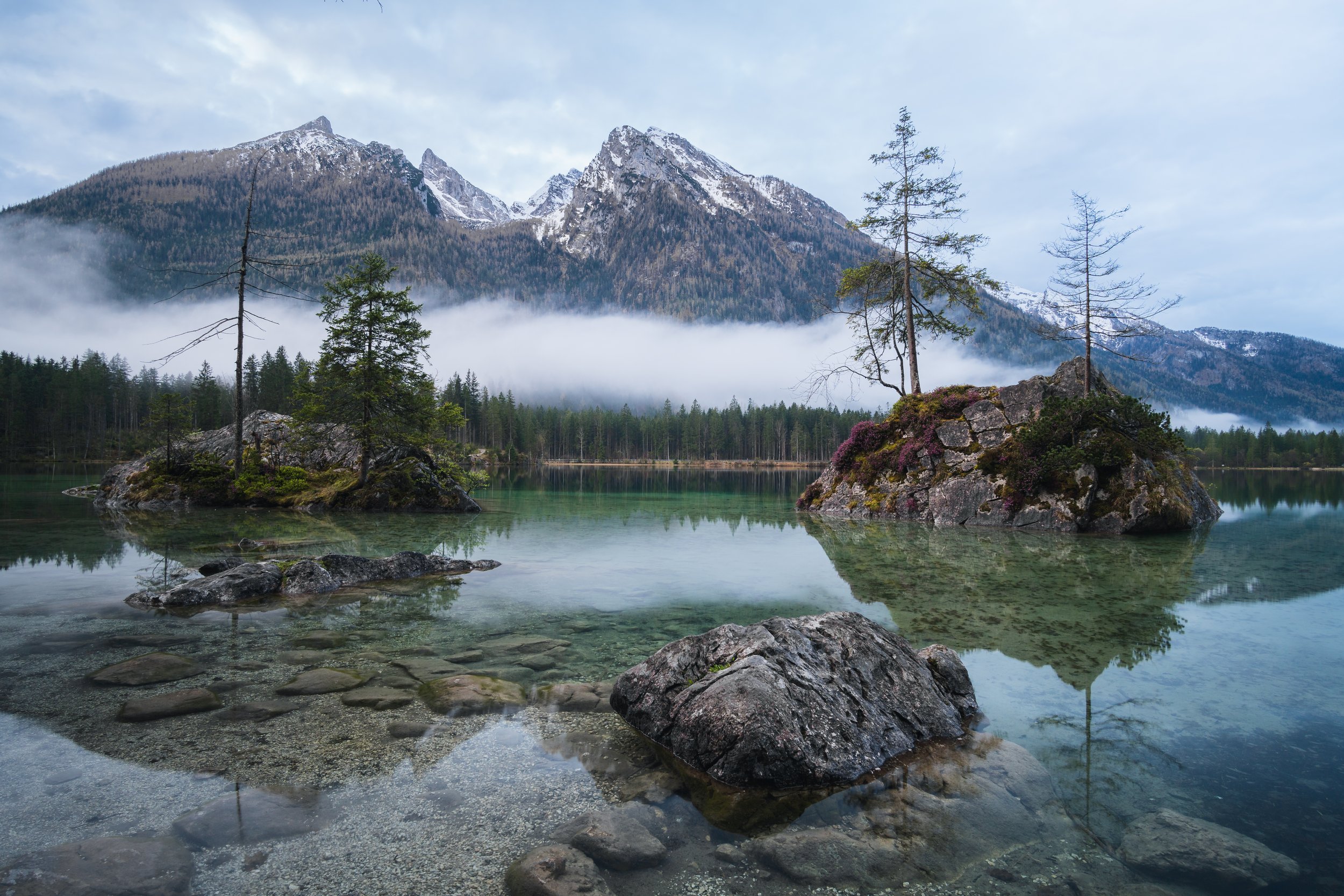 Scenic mountain lake with two small rocky islands featuring trees and shrubs, surrounded by a forest, with snow-capped mountains in the background and mist in the sky.