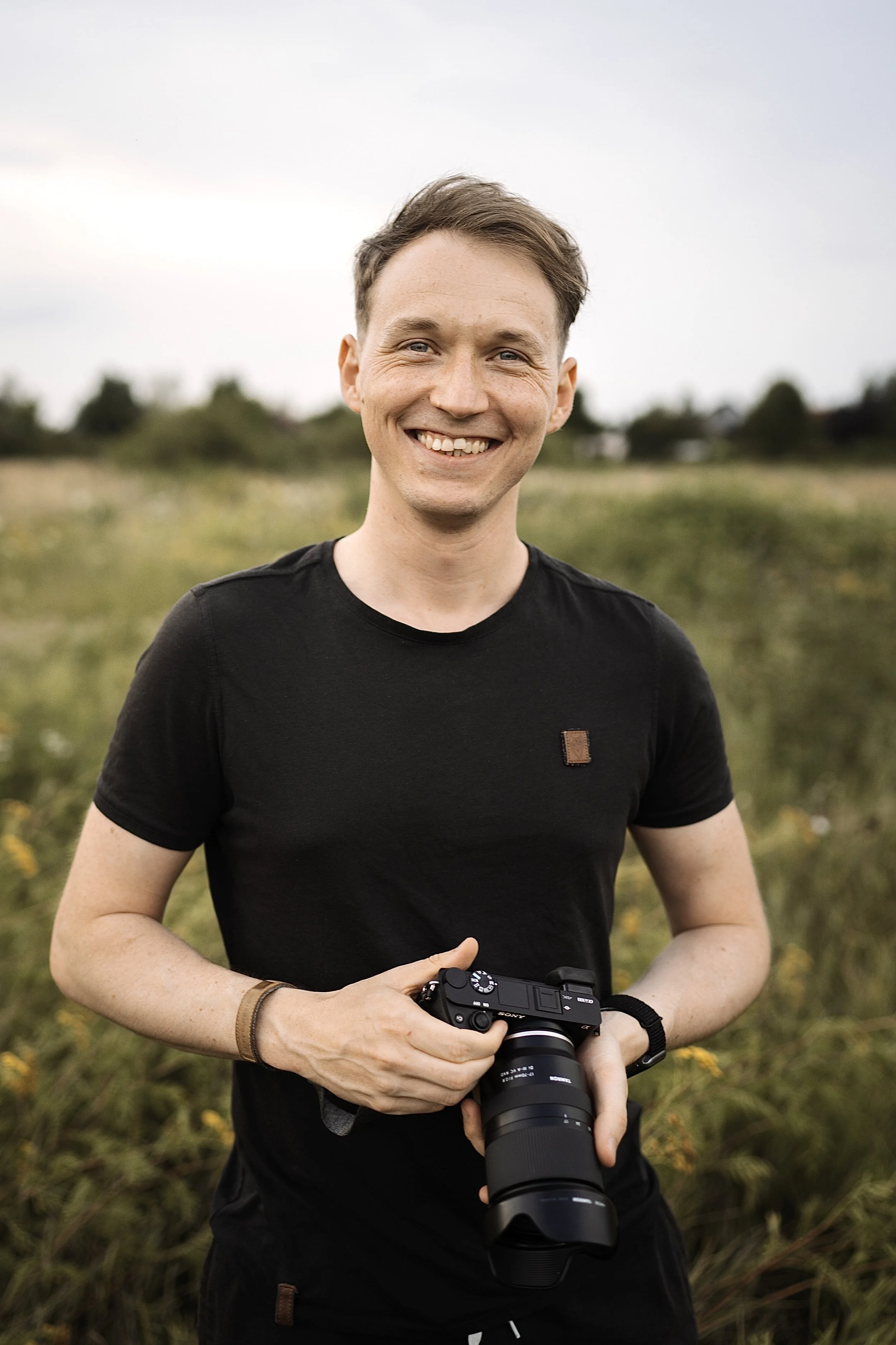 A young man smiling outdoors in a field, holding a camera with a telephoto lens.