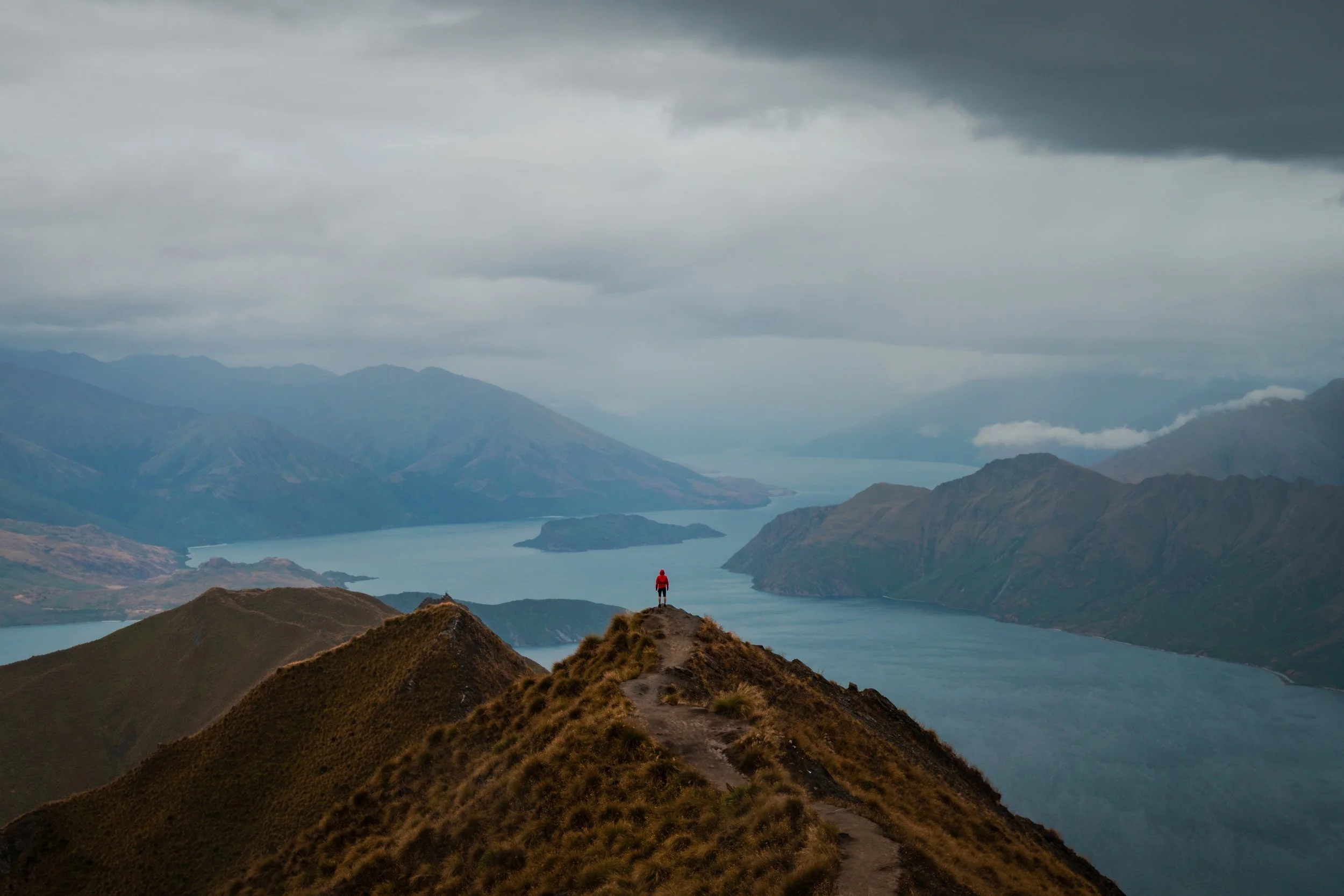 A person standing at the top of a mountain ridge overlooking a large lake and mountain range, under cloudy sky.