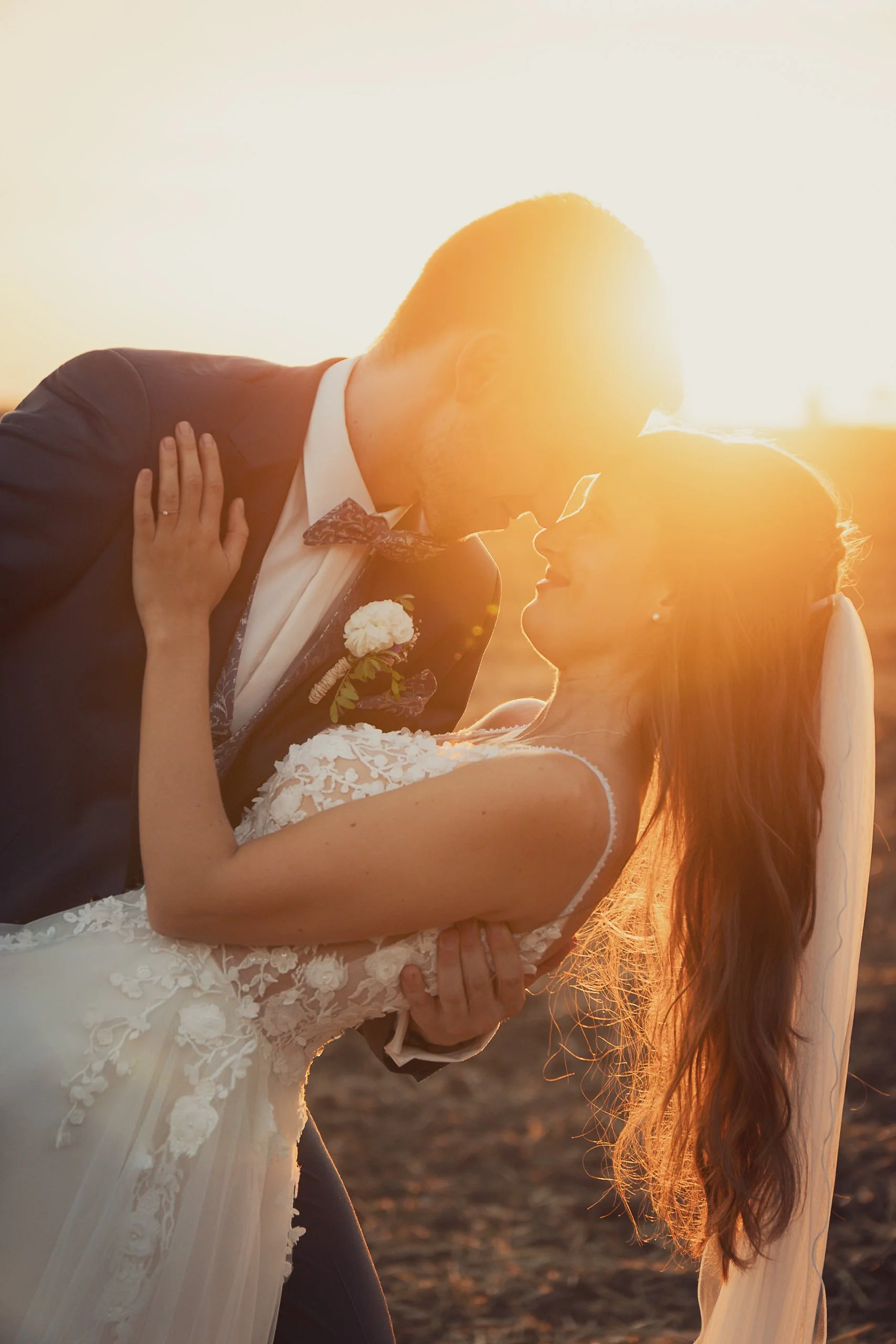 Groom kissing bride during sunset at wedding, bride in white lace dress, groom in dark suit with bow tie, outdoor setting.