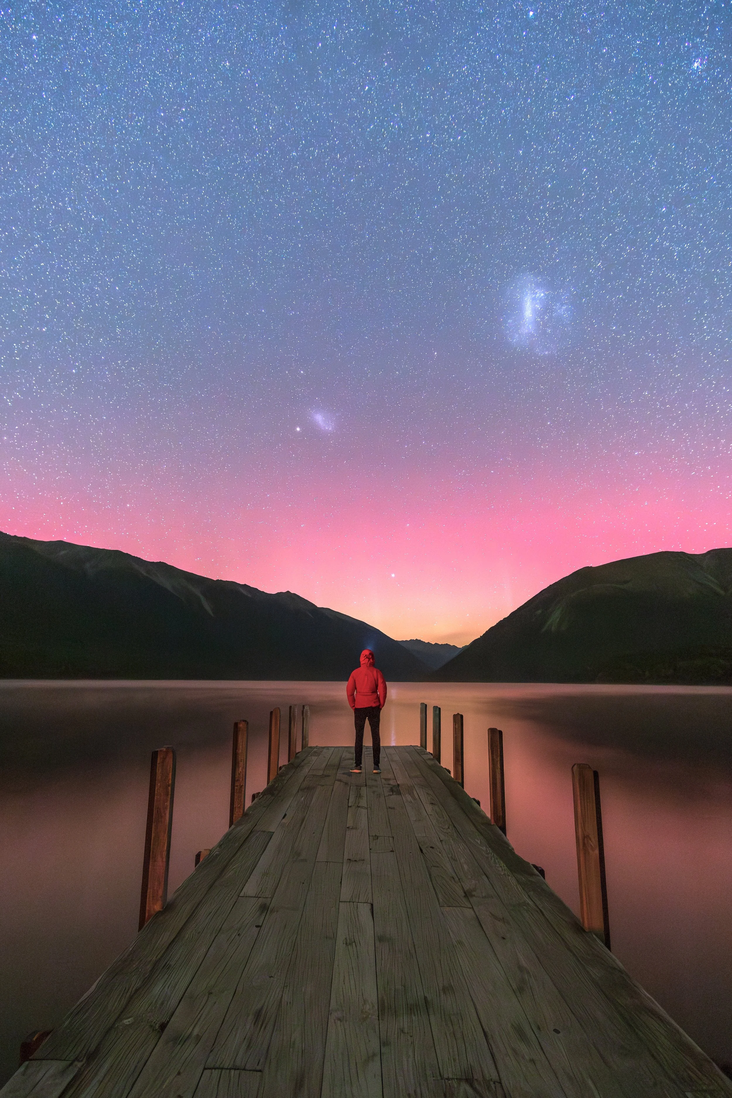 Person in red jacket standing on a wooden dock overlooking a lake at night with a starry sky and the Milky Way galaxy visible above.