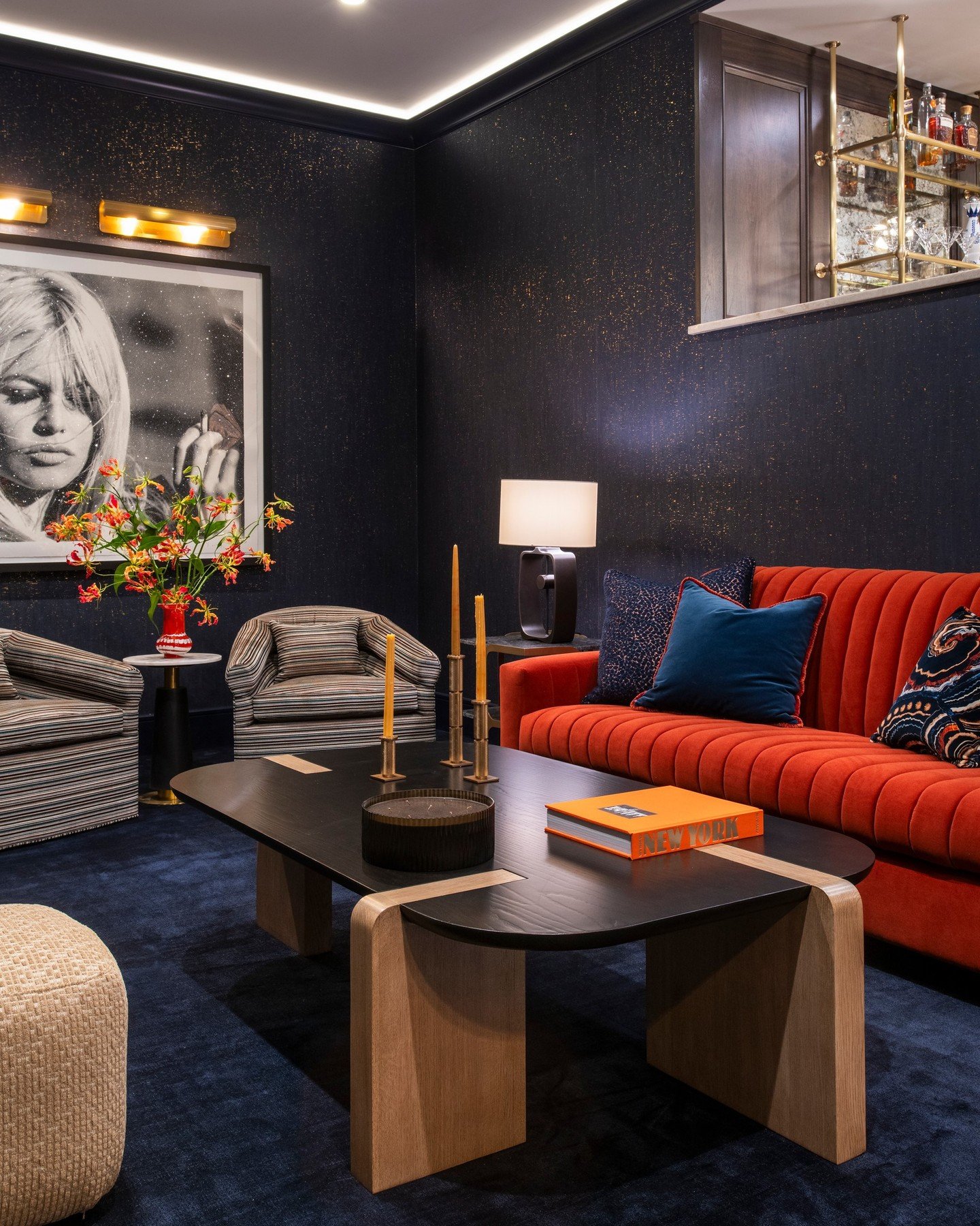 A bold mix of color and texture defines this lower-level lounge.
Deep navy walls shimmer with a hint of metallic, setting the stage for a diamond-dusted Brigitte Bardot artwork that anchors the room.
Vibrant velvet, sculptural furnishings, and moody 