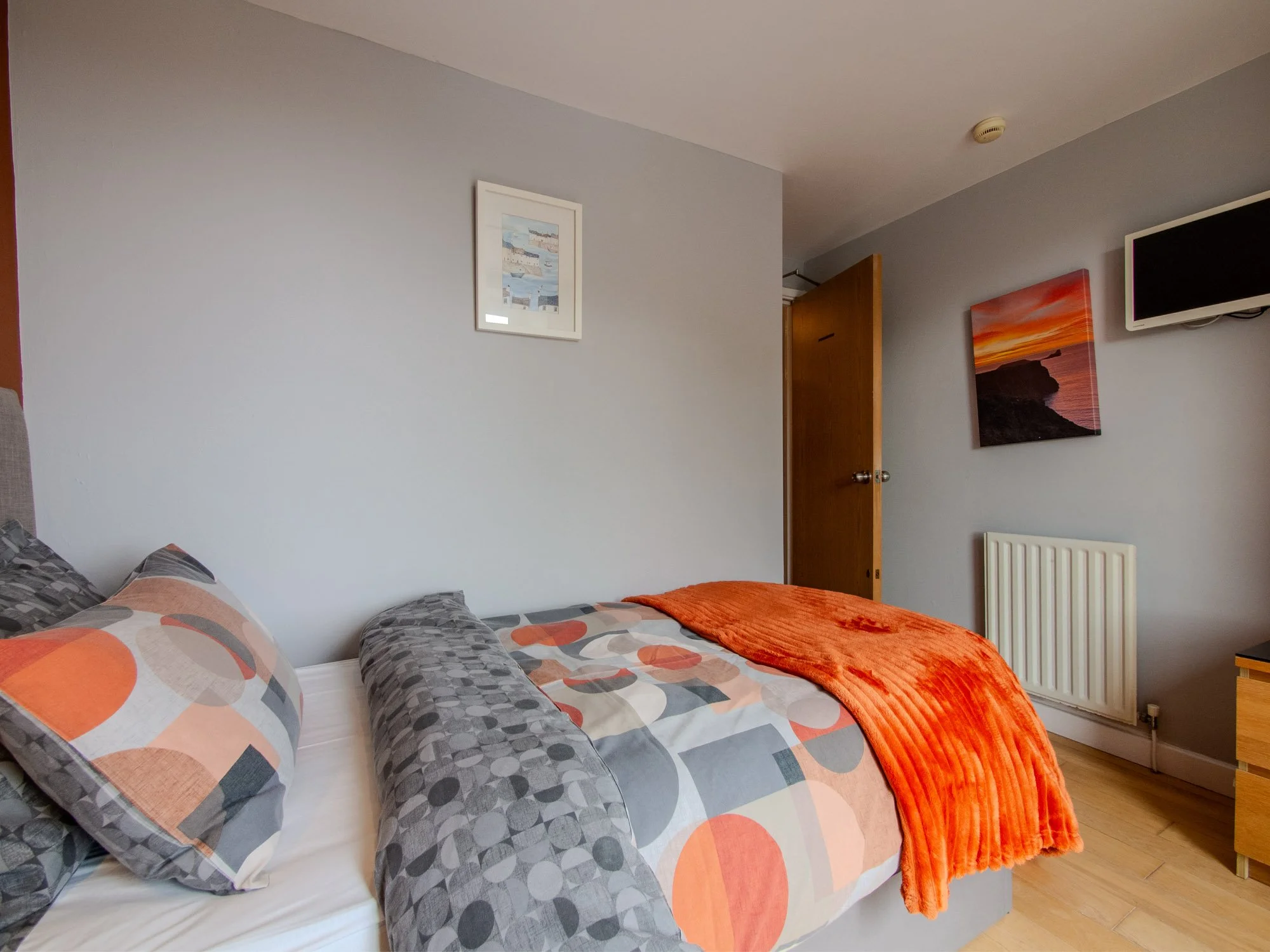A bedroom features a bed with geometric-patterned pillows and a gray and orange bedspread. An orange blanket is draped over the bed. The wall has two framed artworks, one depicting boats and the other a sunset scene. There is a wooden door slightly a