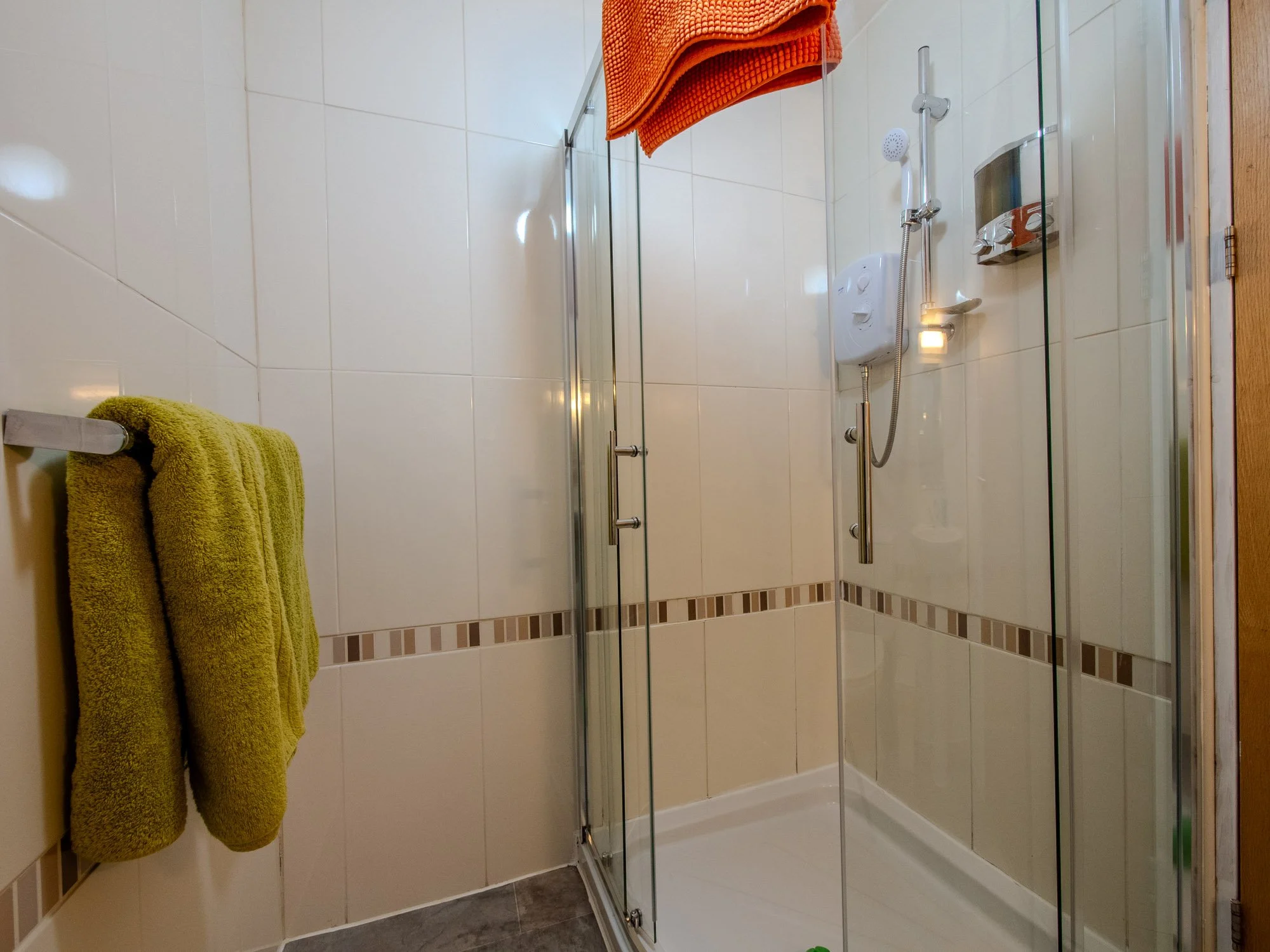 A walk-in shower with glass door, a handheld showerhead, white tiles, and an orange and yellow towel hanging on a rail.