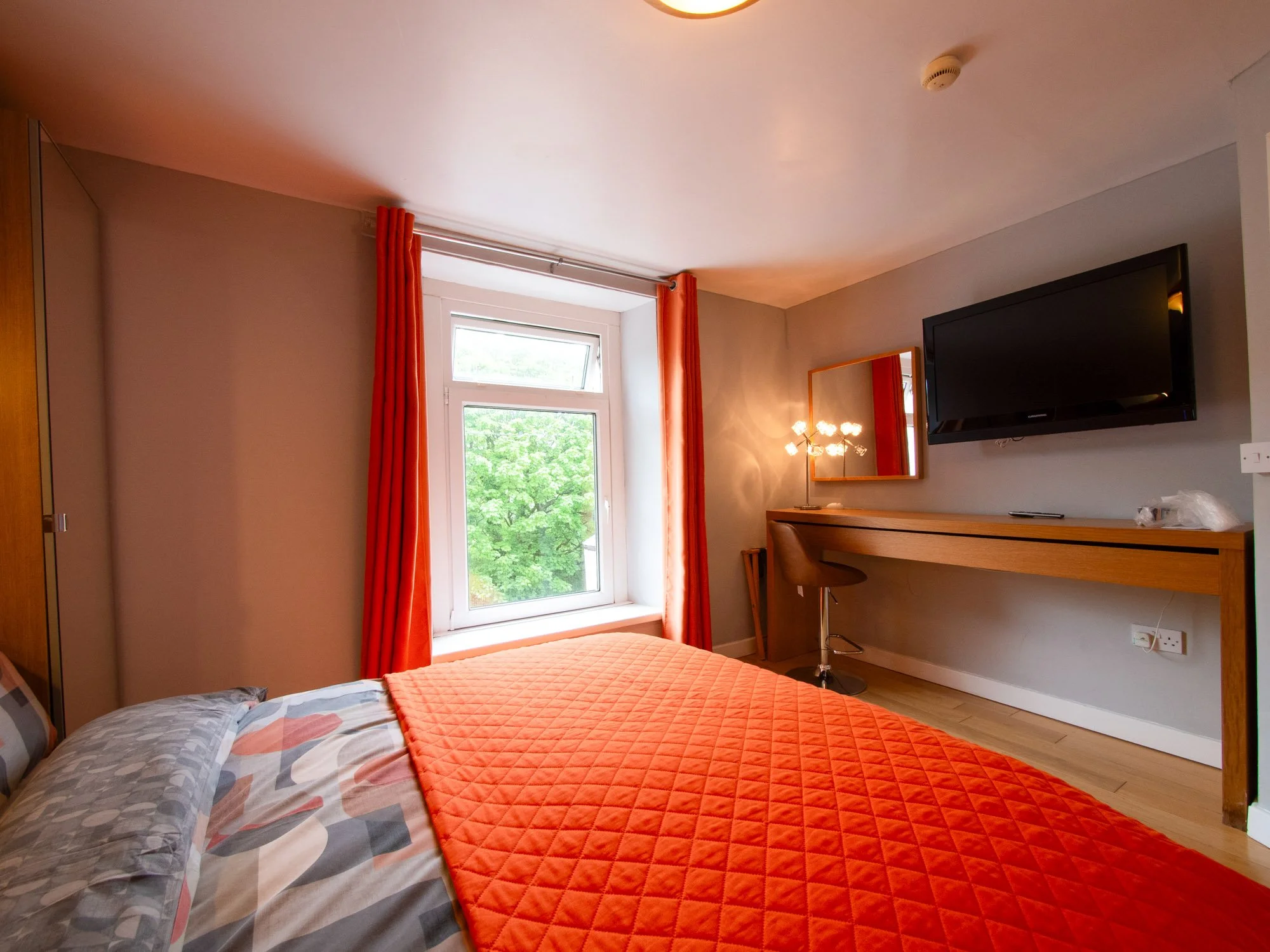 A bedroom with a bed featuring a quilted orange coverlet and patterned pillows, a window with orange curtains letting in natural light, a wall-mounted flat-screen TV, a wooden desk with a chair, a mirror, and a modern light fixture.
