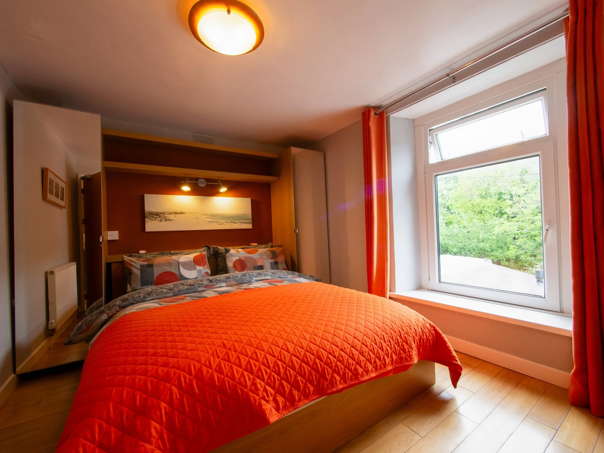 A cozy bedroom with a wooden bed, zebra print pillows, a bright orange quilt, and a large window with orange curtains showing green trees outside. A painting hangs on the wall, and a ceiling light illuminates the room.